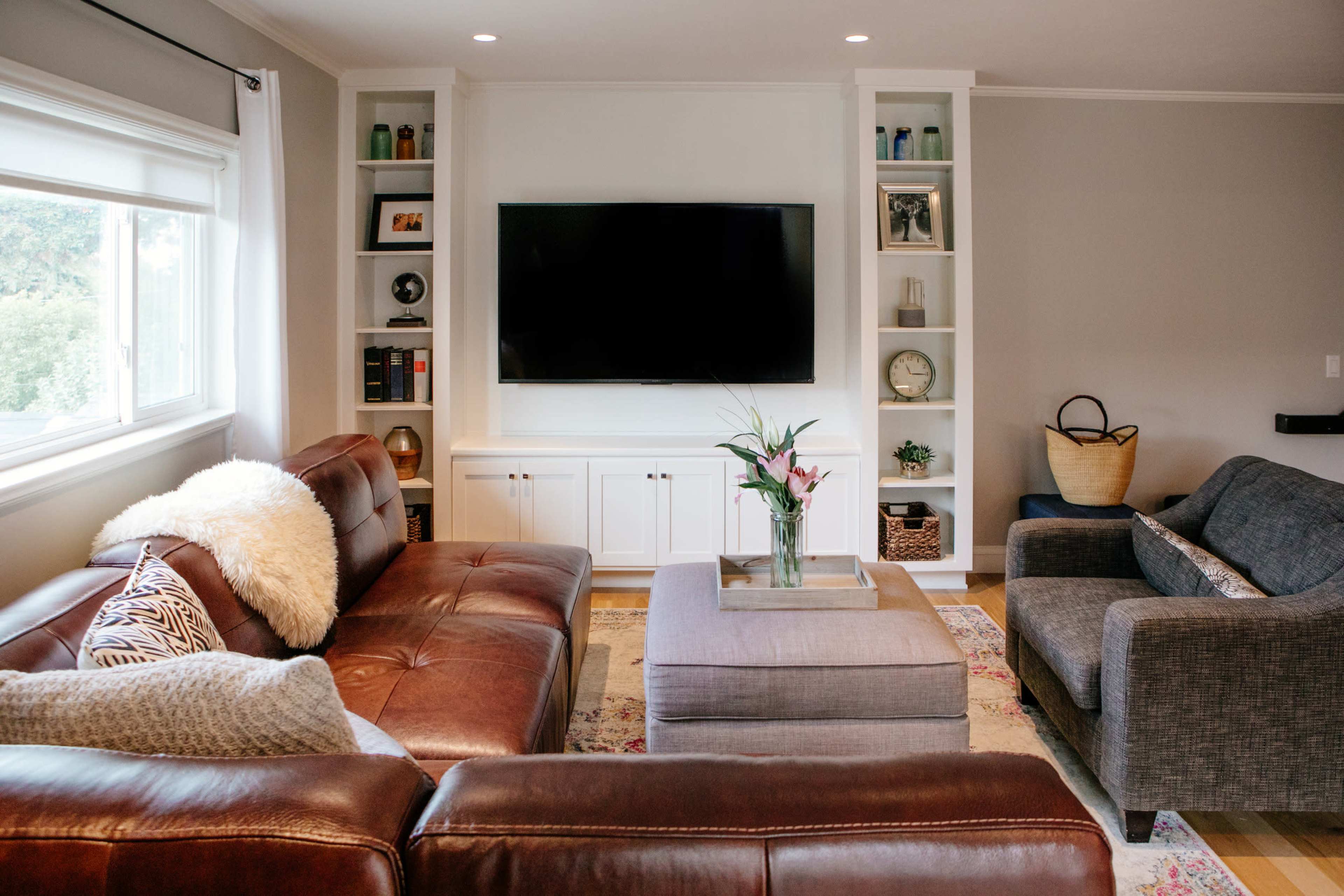 A modern living room features a large flat-screen TV mounted on a white cabinet, with dark leather sofas and a grey ottoman at the center.