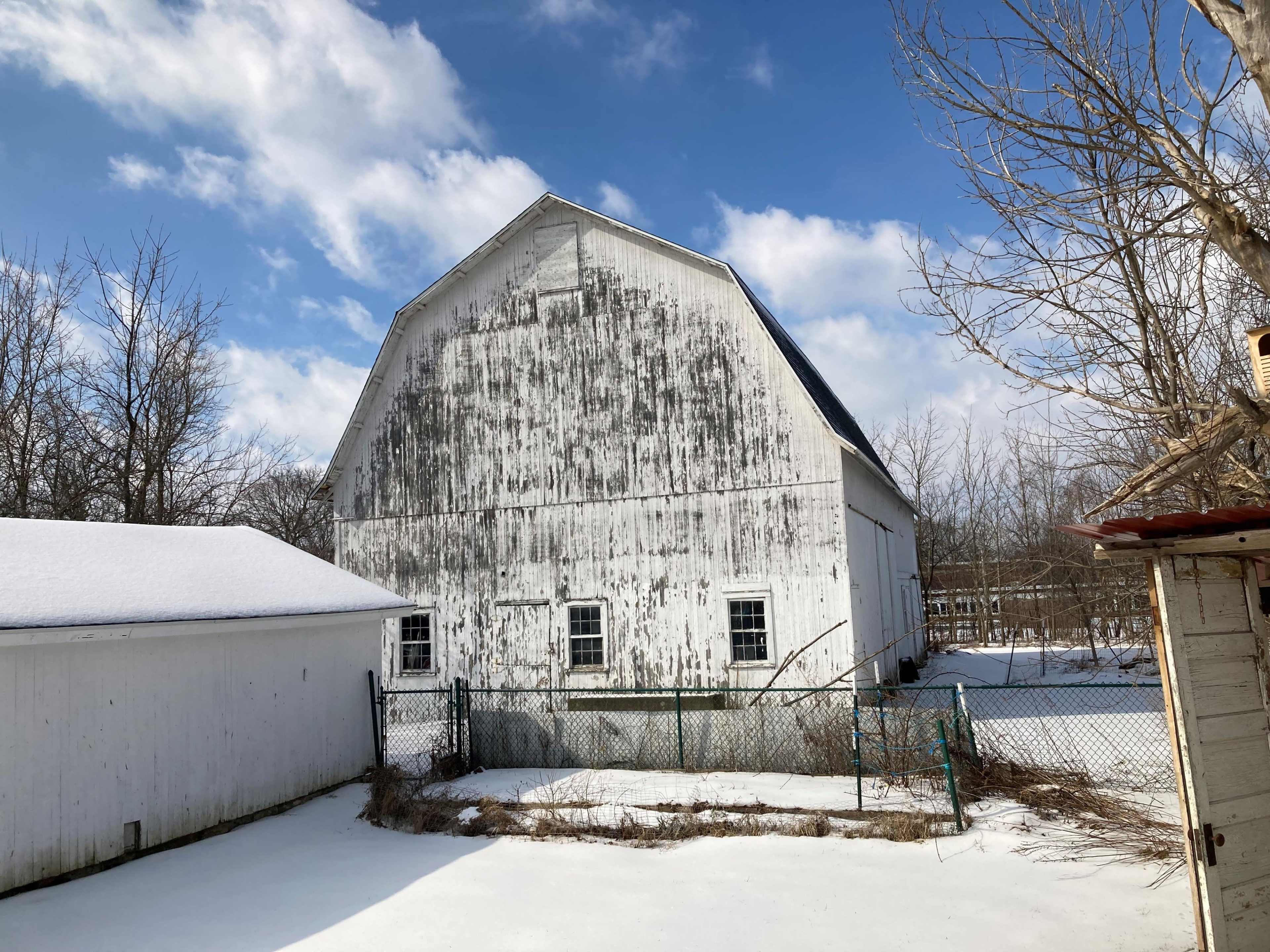 A weathered white barn with a sloped roof, surrounded by a snowy landscape and bare trees under a partly cloudy sky.