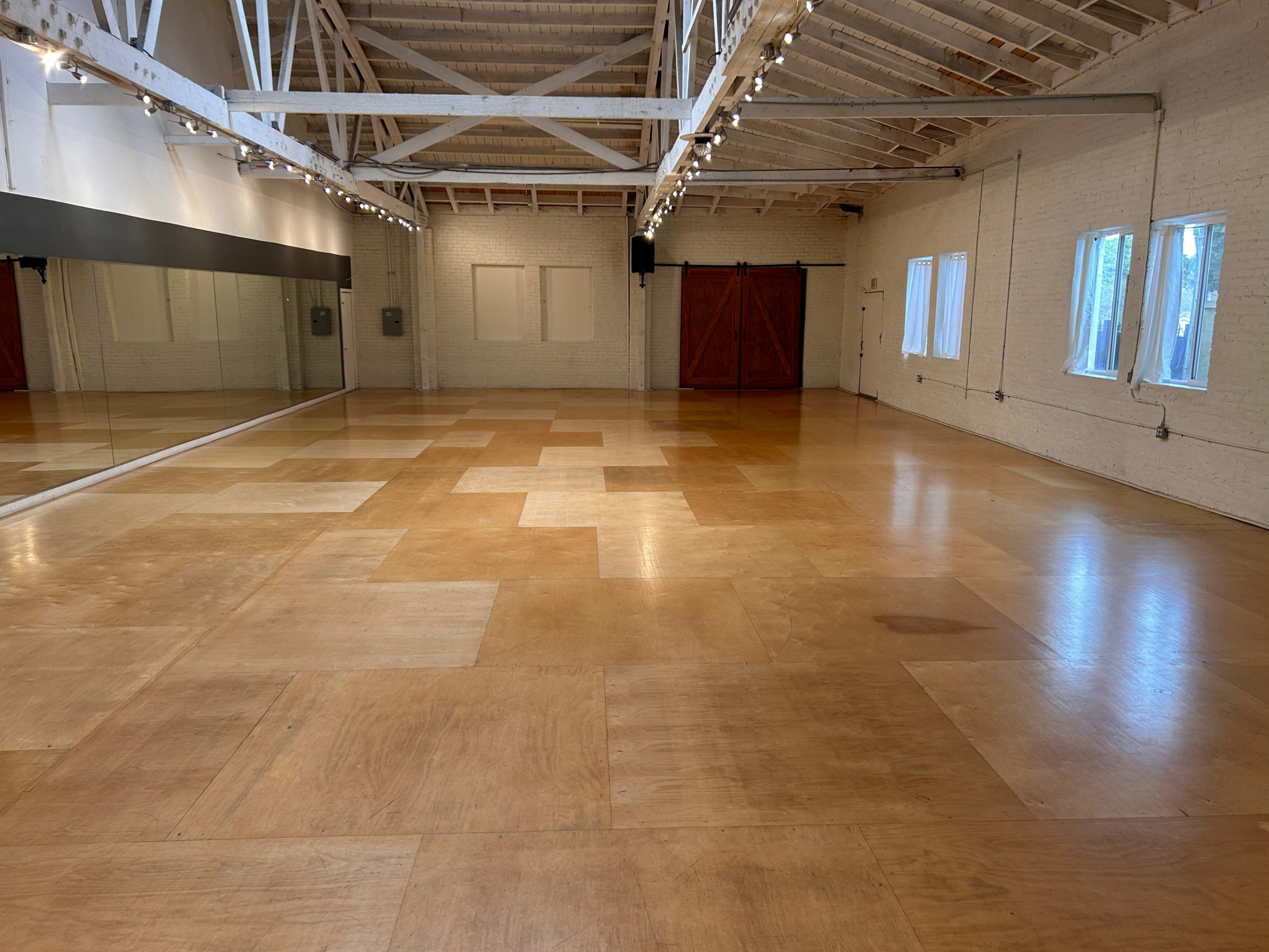 A spacious, empty room with wooden flooring, large windows, and mirrored walls.