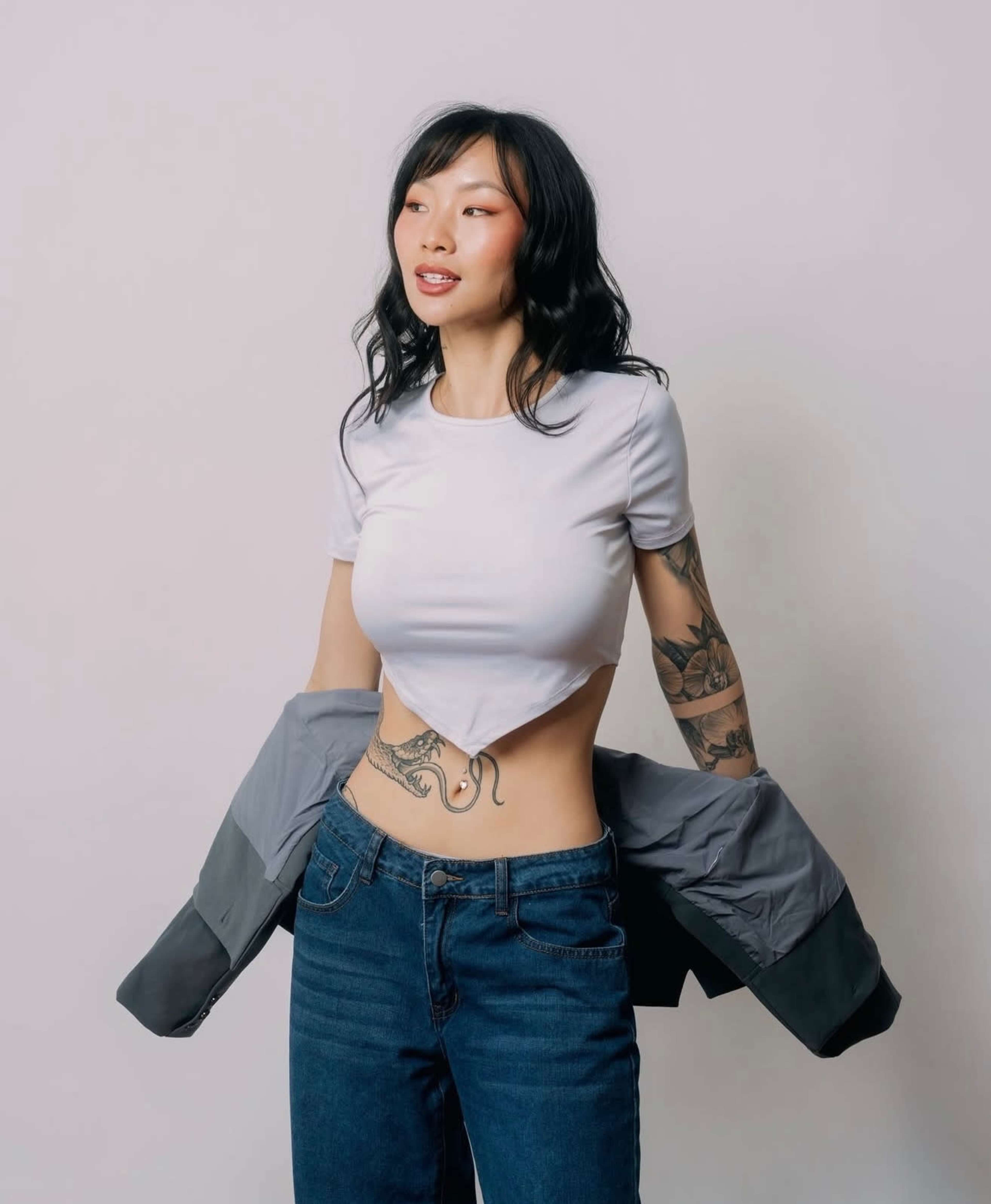 A woman with tattoos poses in a cropped white t-shirt and blue jeans while holding a gray jacket.
