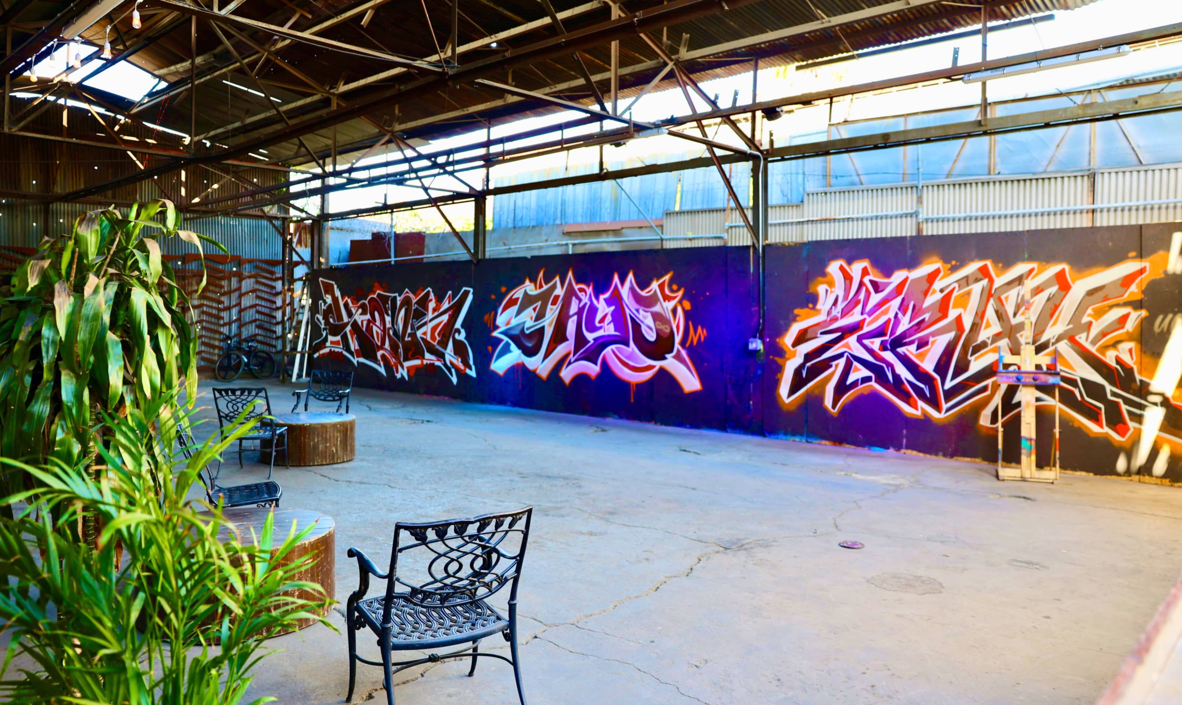 The image shows an industrial space featuring vibrant graffiti on one wall and metal chairs arranged on the concrete floor.