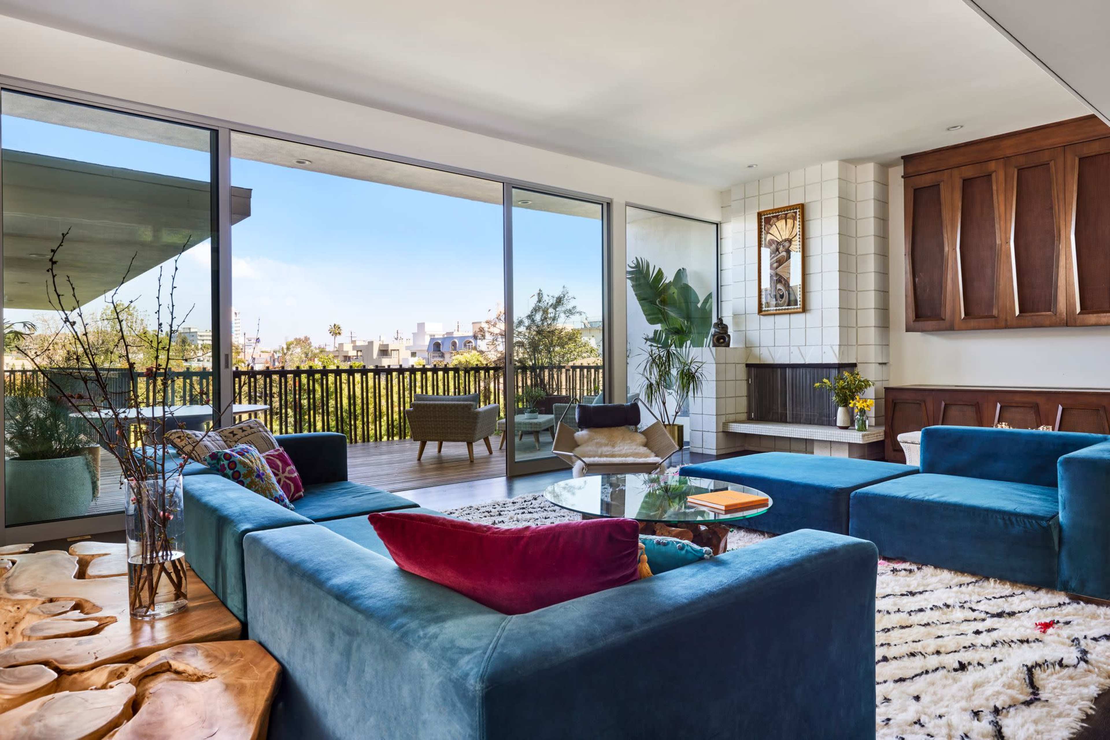 The image shows a modern living room featuring a blue sectional sofa, a coffee table, large windows providing a view of an outdoor balcony, and various decorative elements.