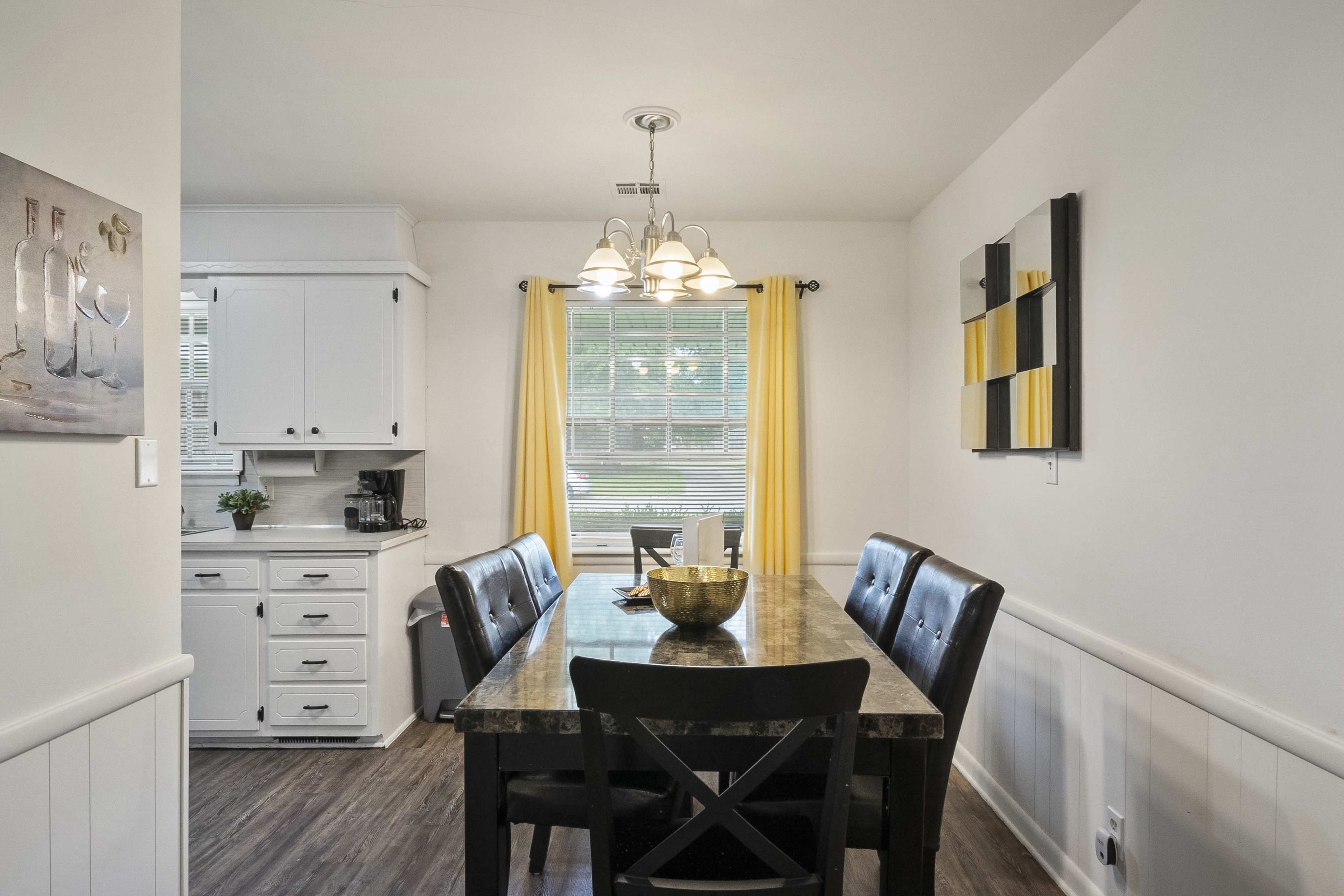 A dining area features a black table surrounded by four chairs, with yellow curtains framing a window that overlooks a kitchen.
