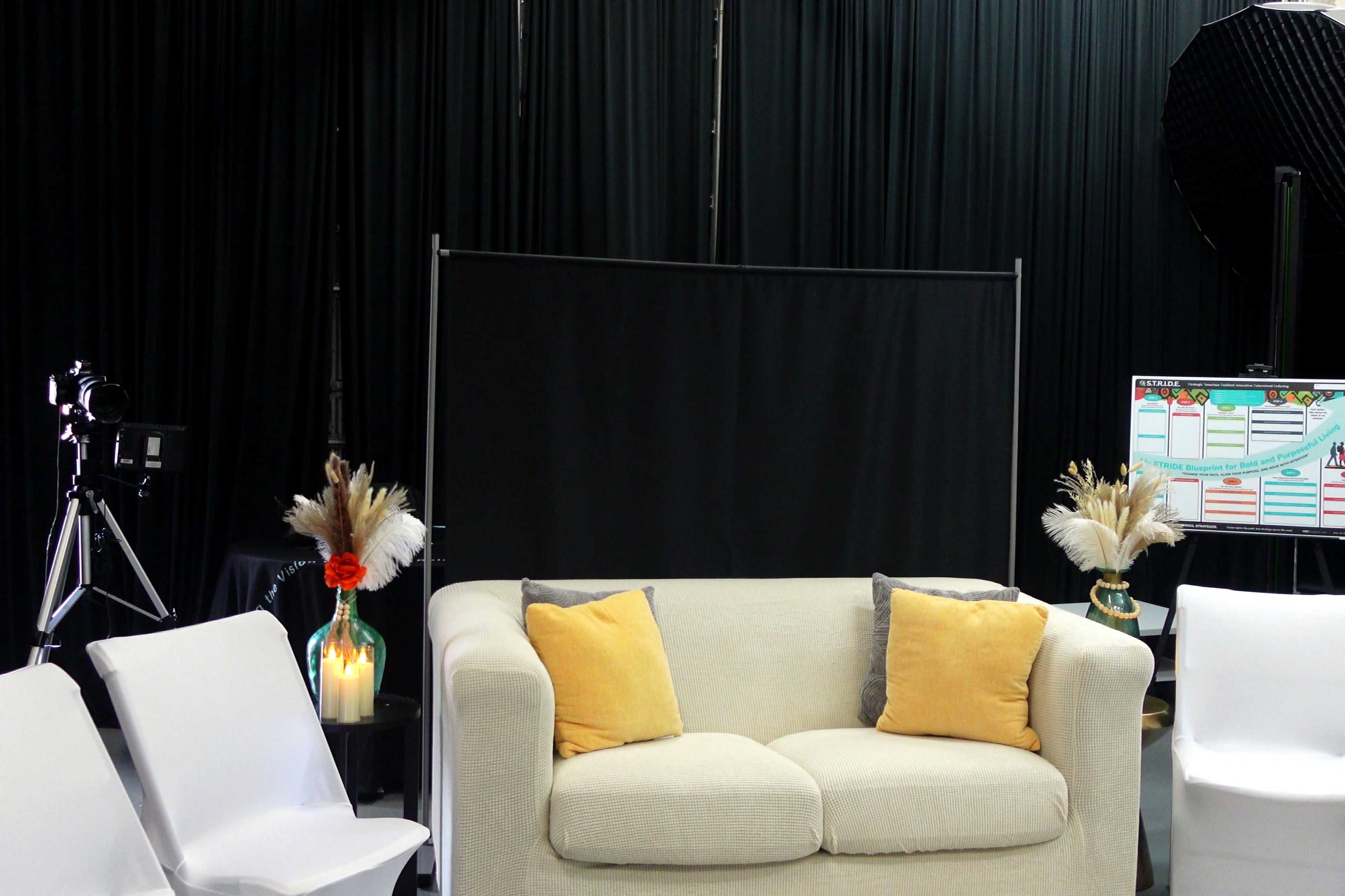The image shows a studio setting with a white couch adorned with yellow and gray pillows, flanked by vases of dried flowers, a black backdrop, and a camera setup.