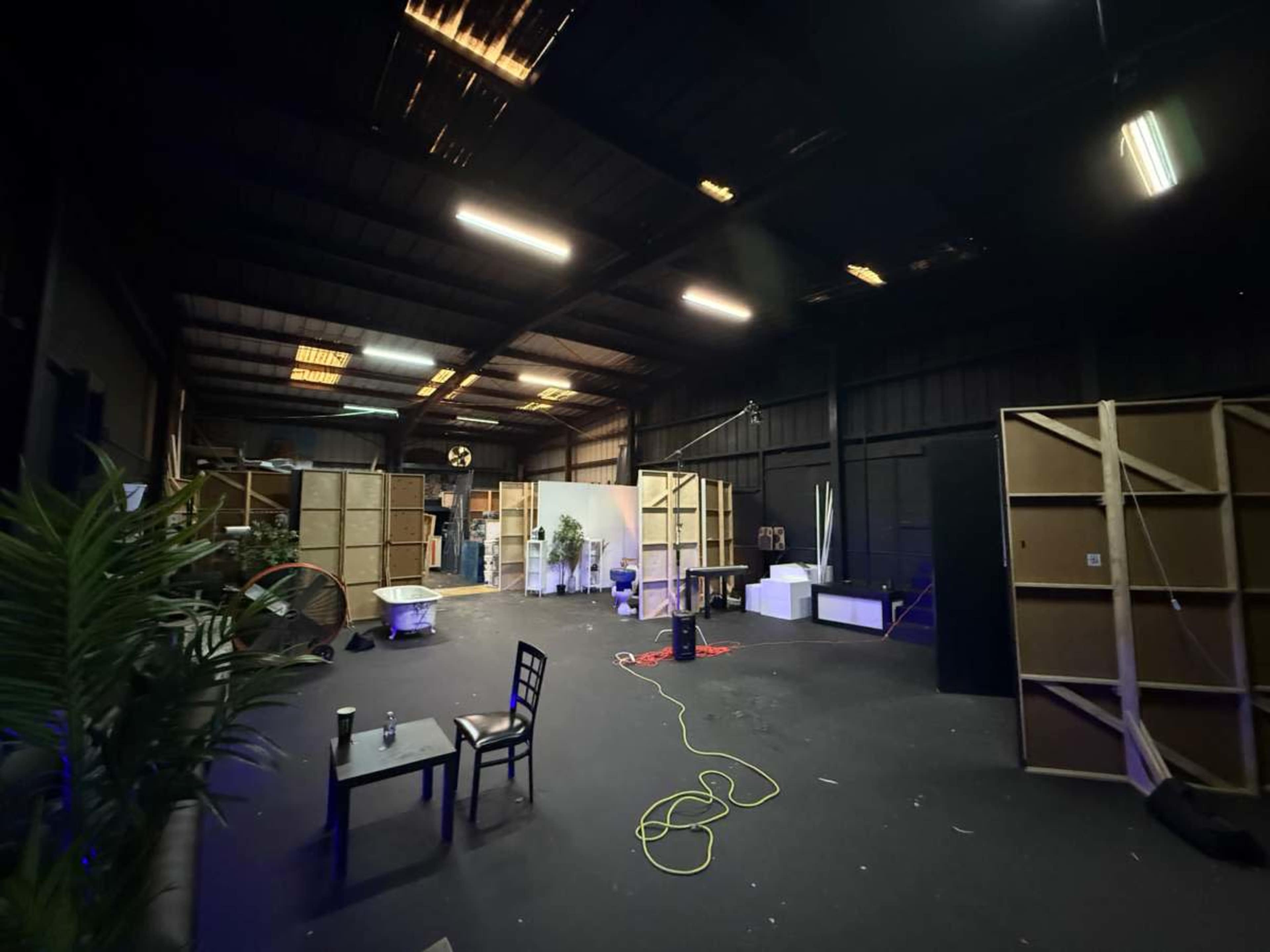 The image shows an industrial space with a dark floor and walls, featuring wooden frames, a few plants, and scattered furniture in a partially lit environment.