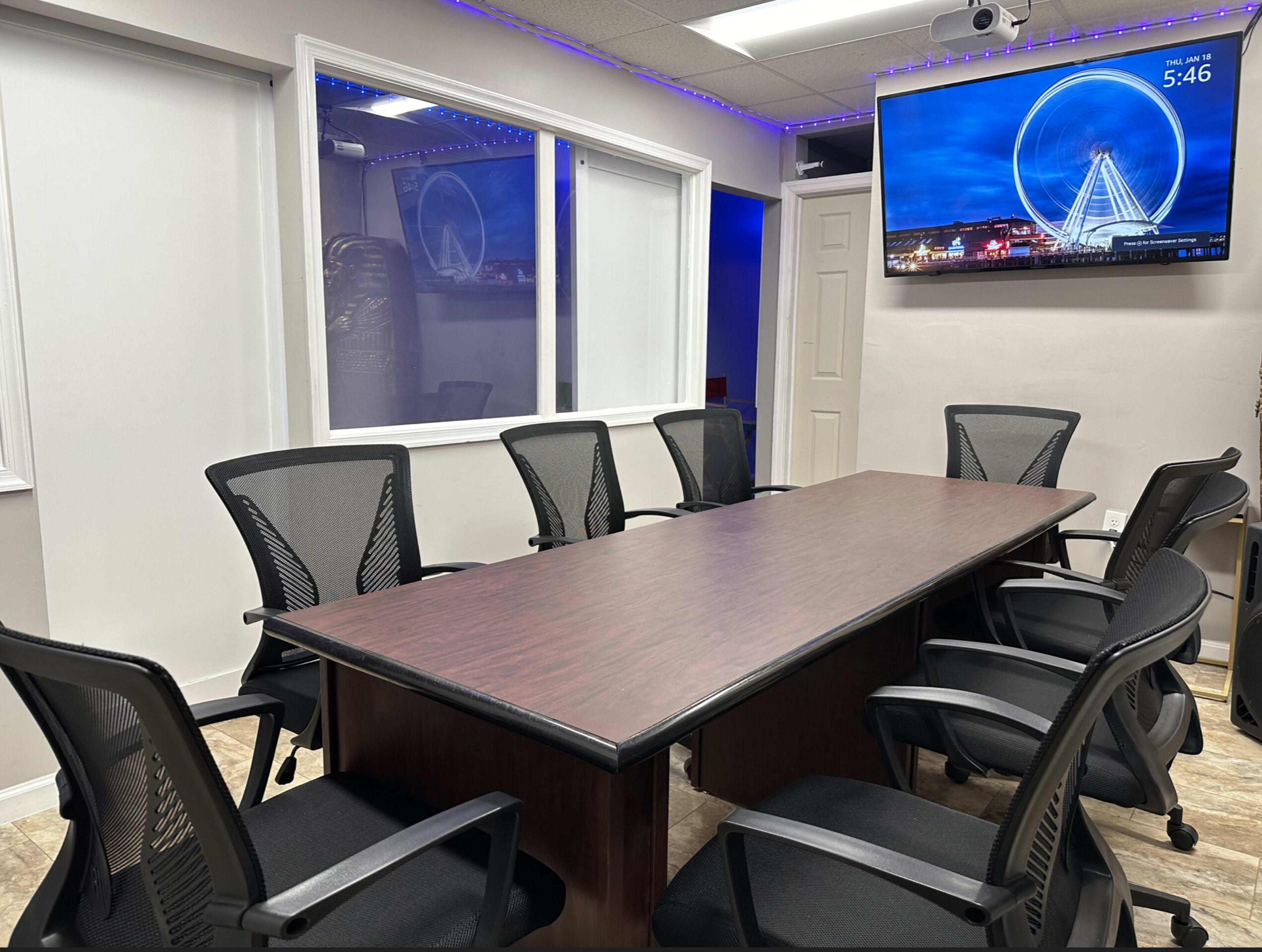A conference room with a long brown table surrounded by six black mesh chairs and a large wall-mounted screen displaying a wheel structure against a blue background.