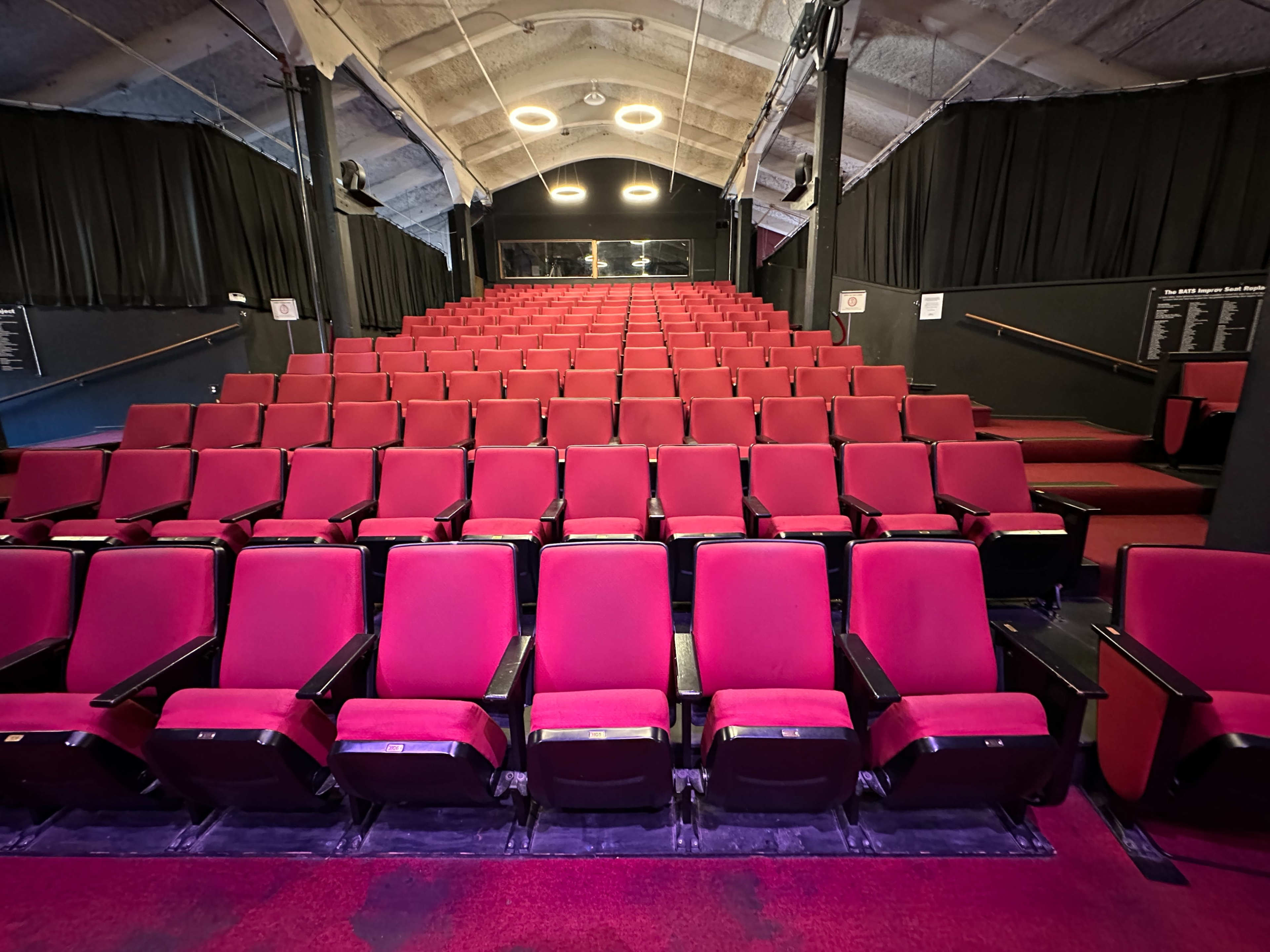 A semi-empty theater with rows of red cushioned seats facing a stage.