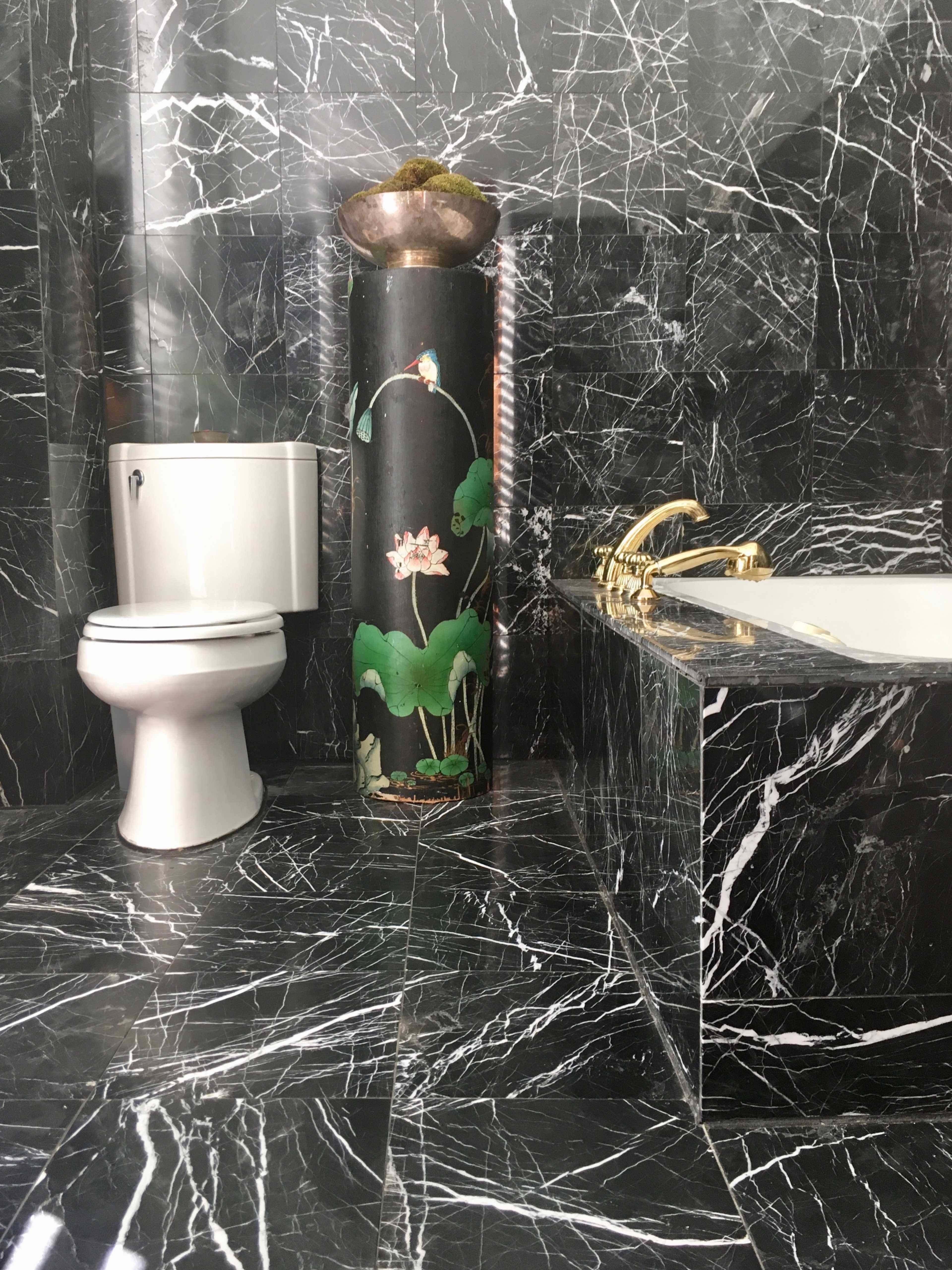 The bathroom features black marble walls and flooring, a white toilet, a sculpted column with a decorative bowl, and a bathtub with gold fixtures.