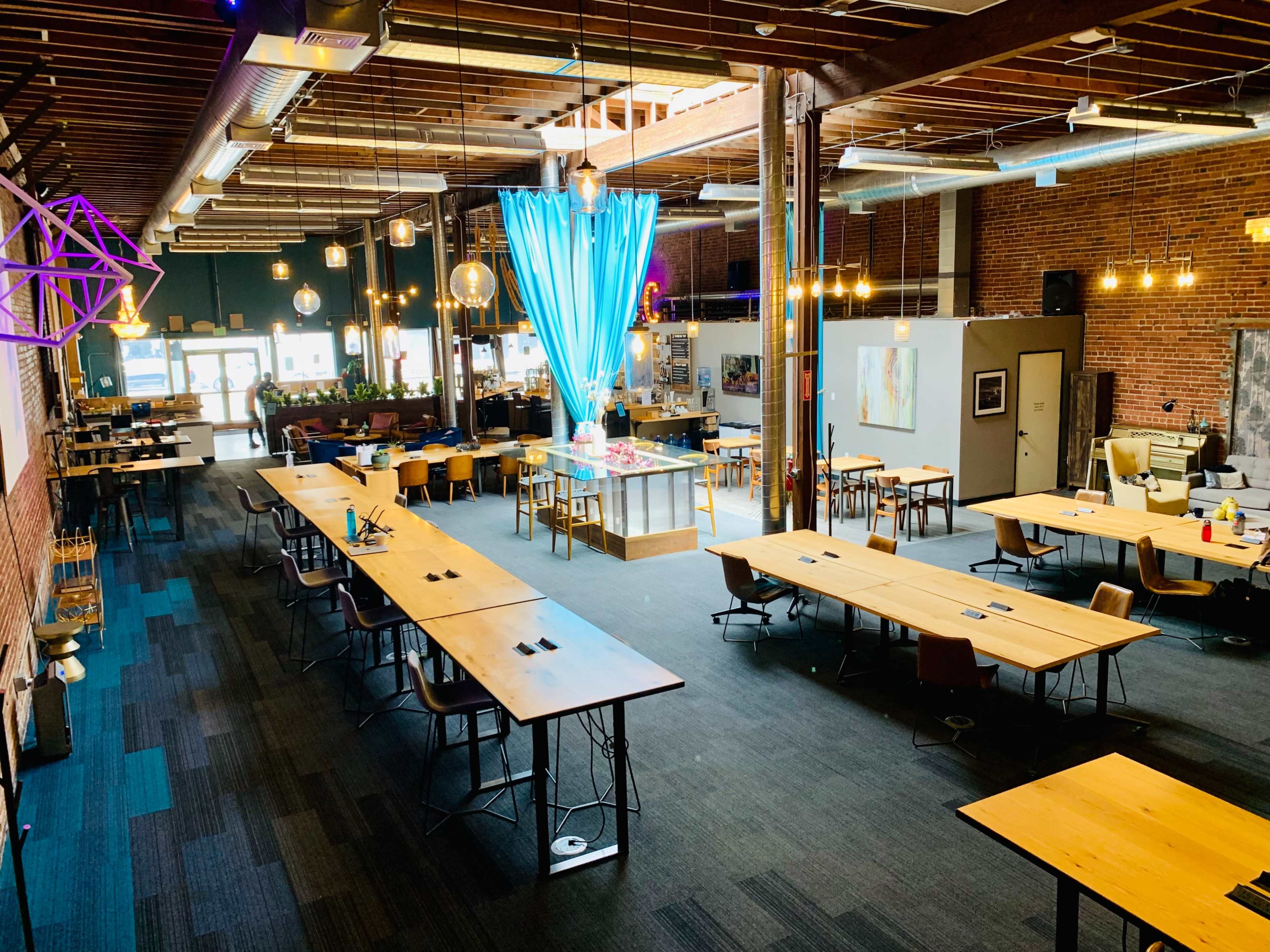 A spacious, modern workspace with multiple wooden tables arranged in rows, bright blue lighting hanging from the ceiling, and exposed brick walls.