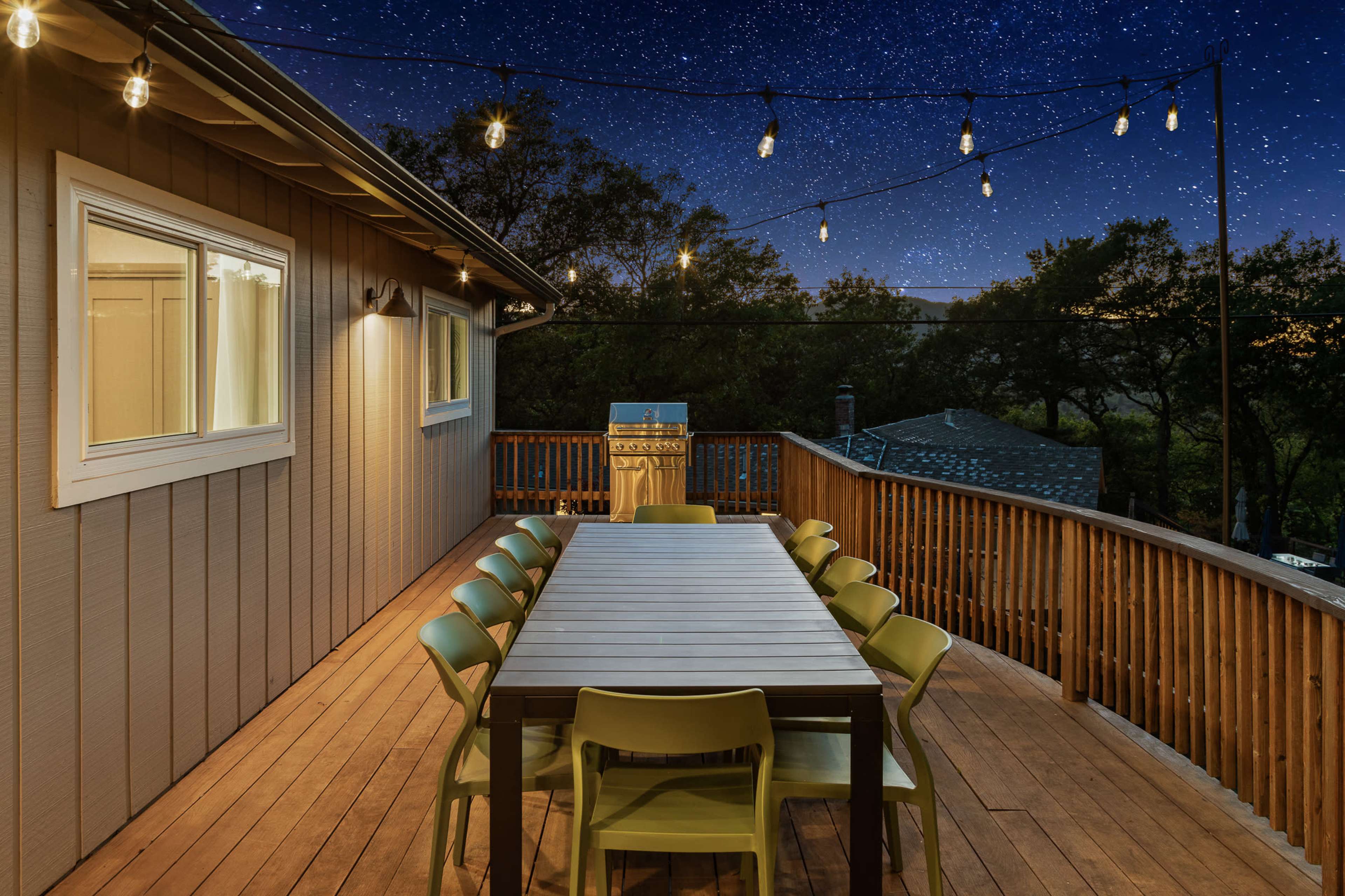 A wooden deck features a long dining table surrounded by green chairs, with string lights overhead and a starry night sky visible in the background.