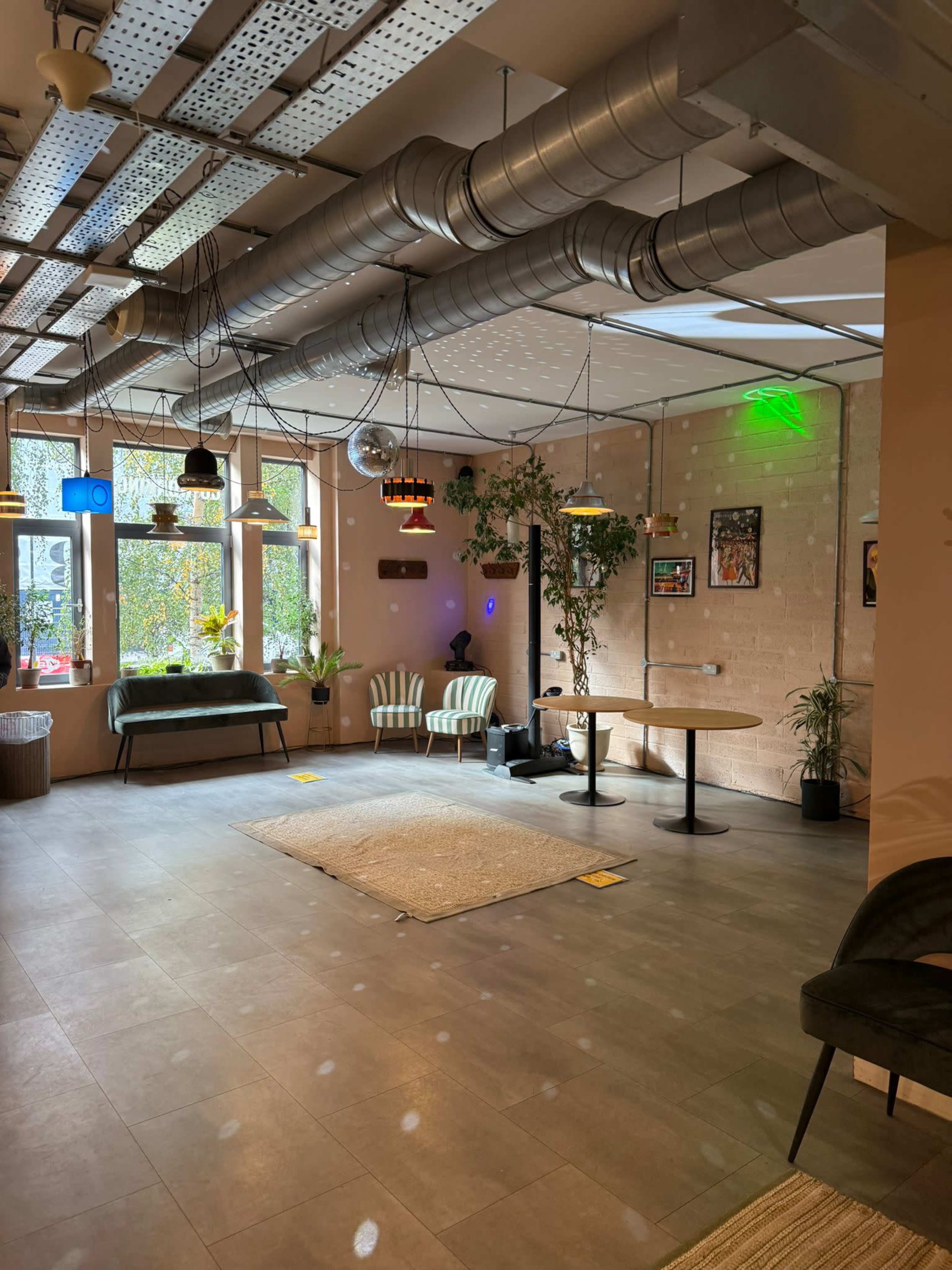 The image depicts a modern, well-lit interior space with stylish seating, decorative plants, and unique light fixtures hanging from the ceiling.