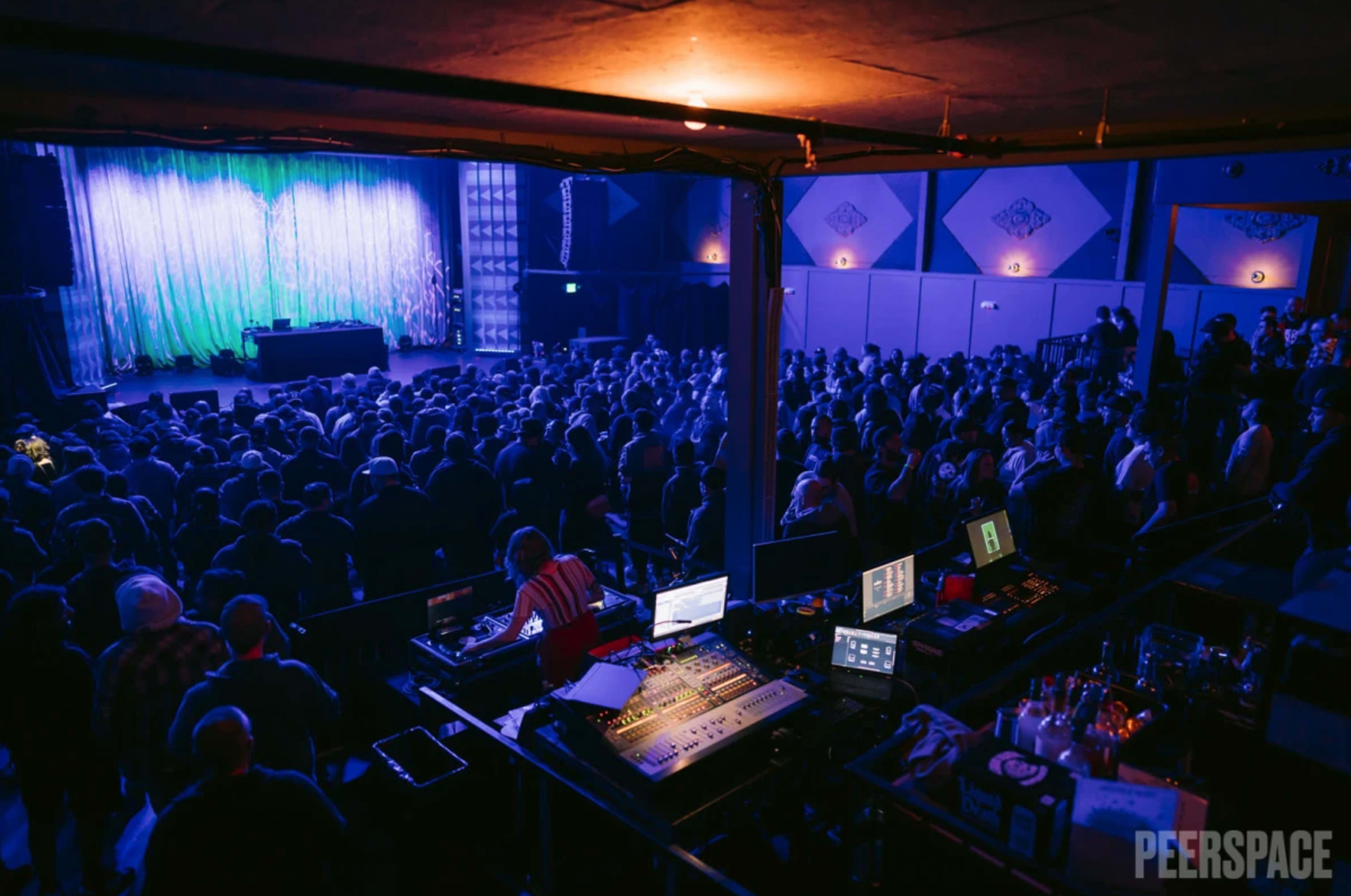 A large crowd is gathered in a dimly lit venue facing a stage with a shimmering backdrop and audio equipment set up in the foreground.