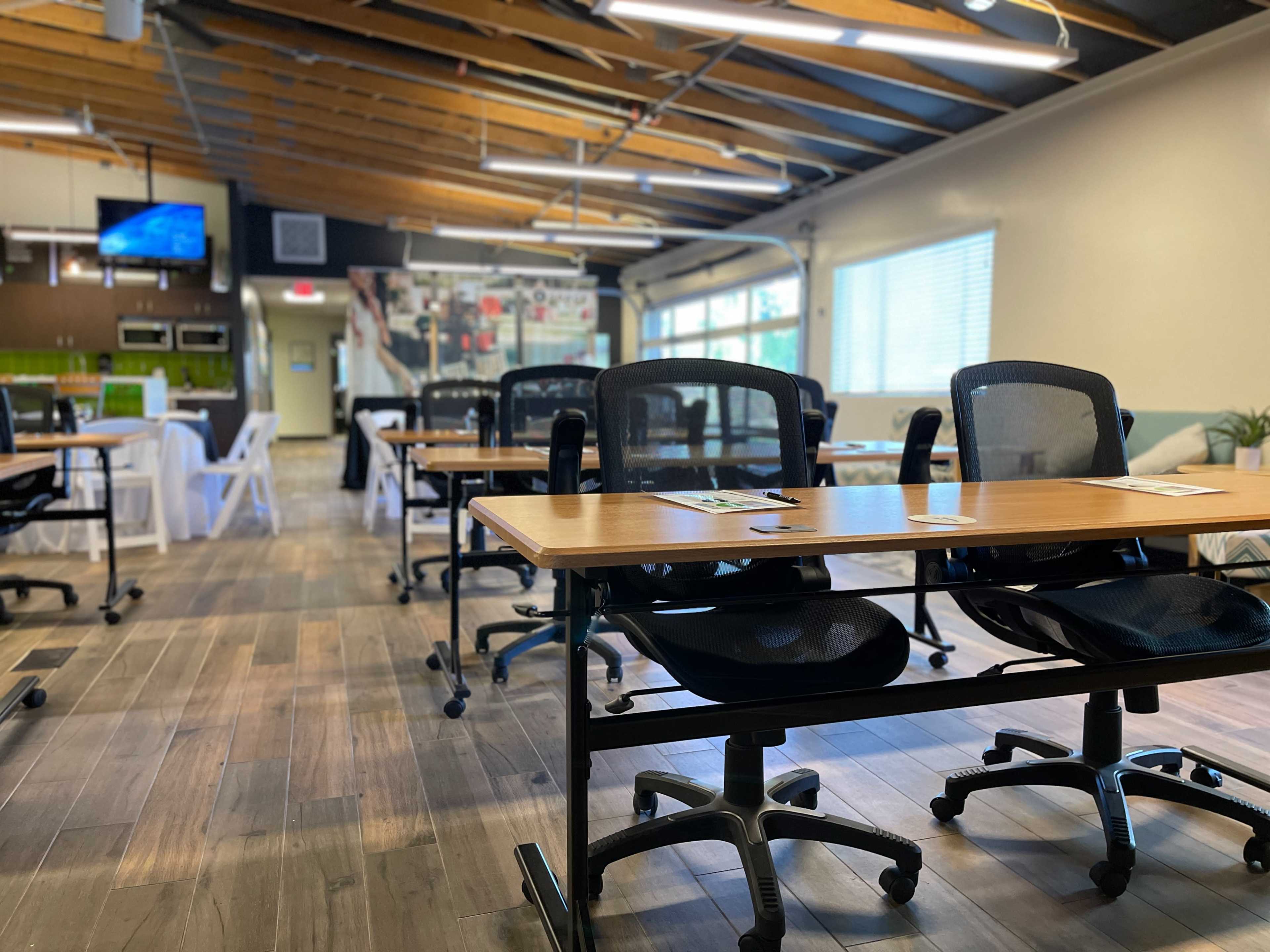 An empty classroom featuring several rows of tables and chairs arranged neatly under a high ceiling with wooden beams.