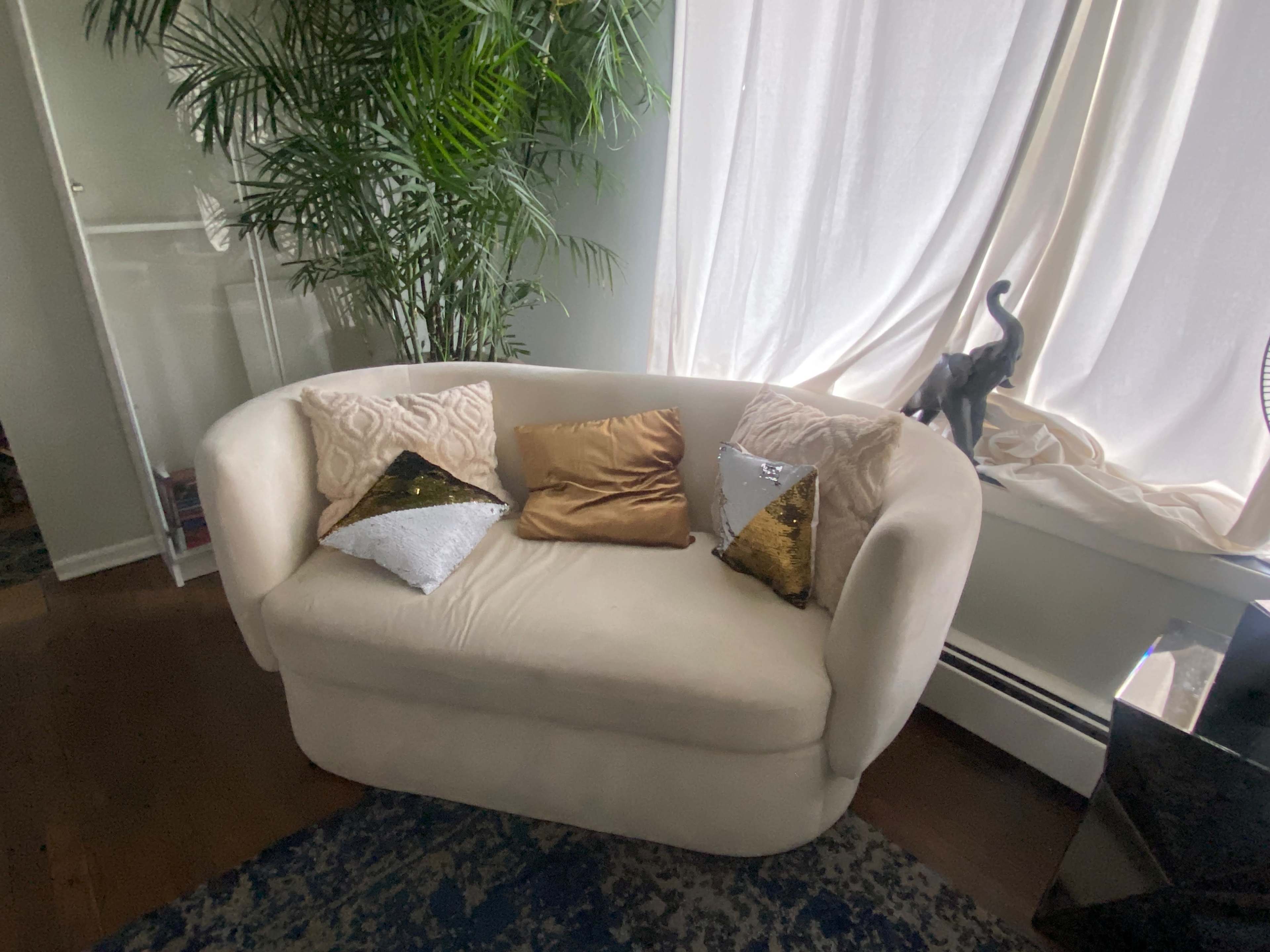 A beige loveseat adorned with decorative pillows in various colors, situated near a window with sheer curtains and a potted plant in the background.