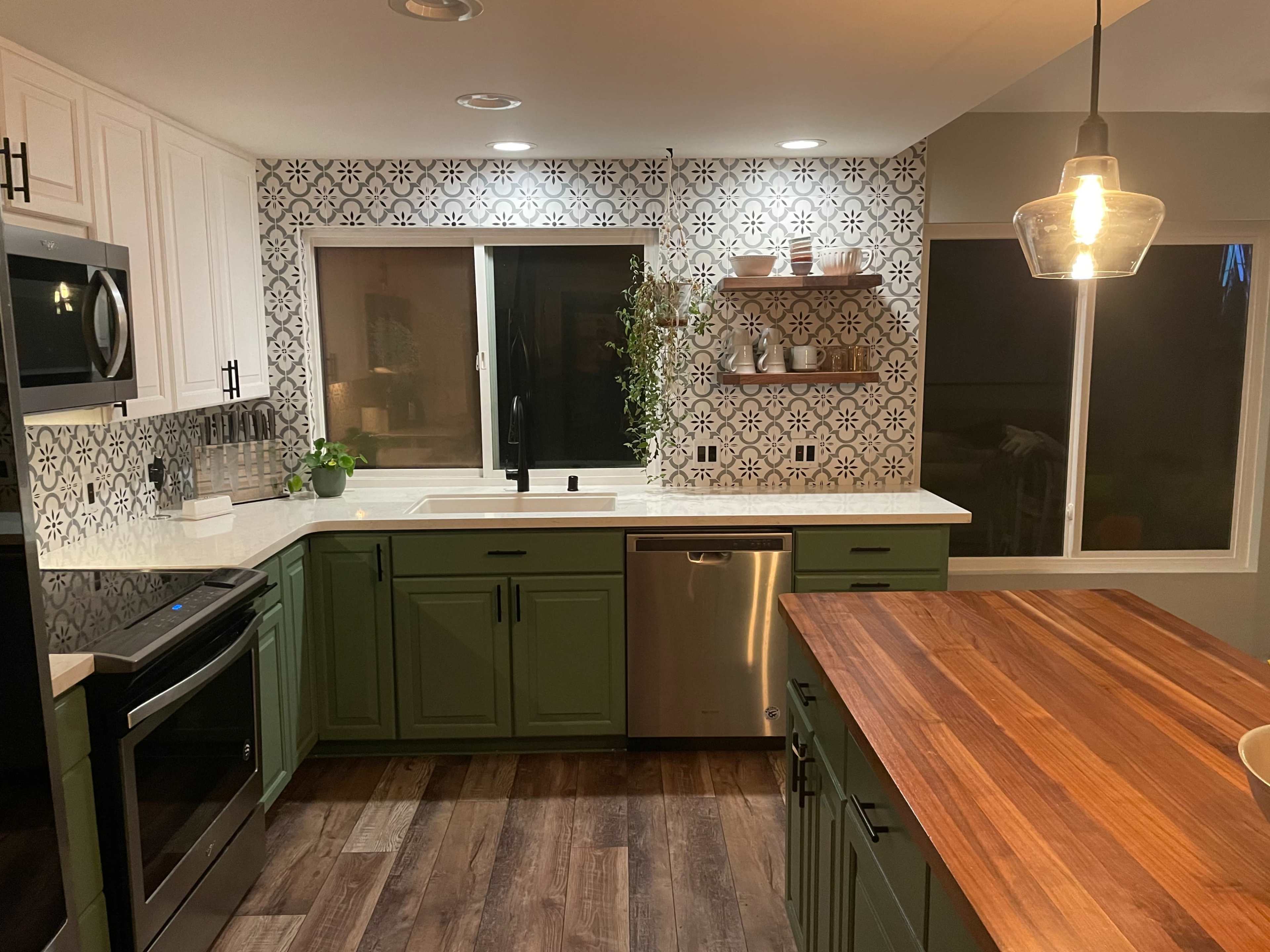 A modern kitchen features green cabinets, a wooden countertop, and patterned tiles on the wall, with a window providing natural light.