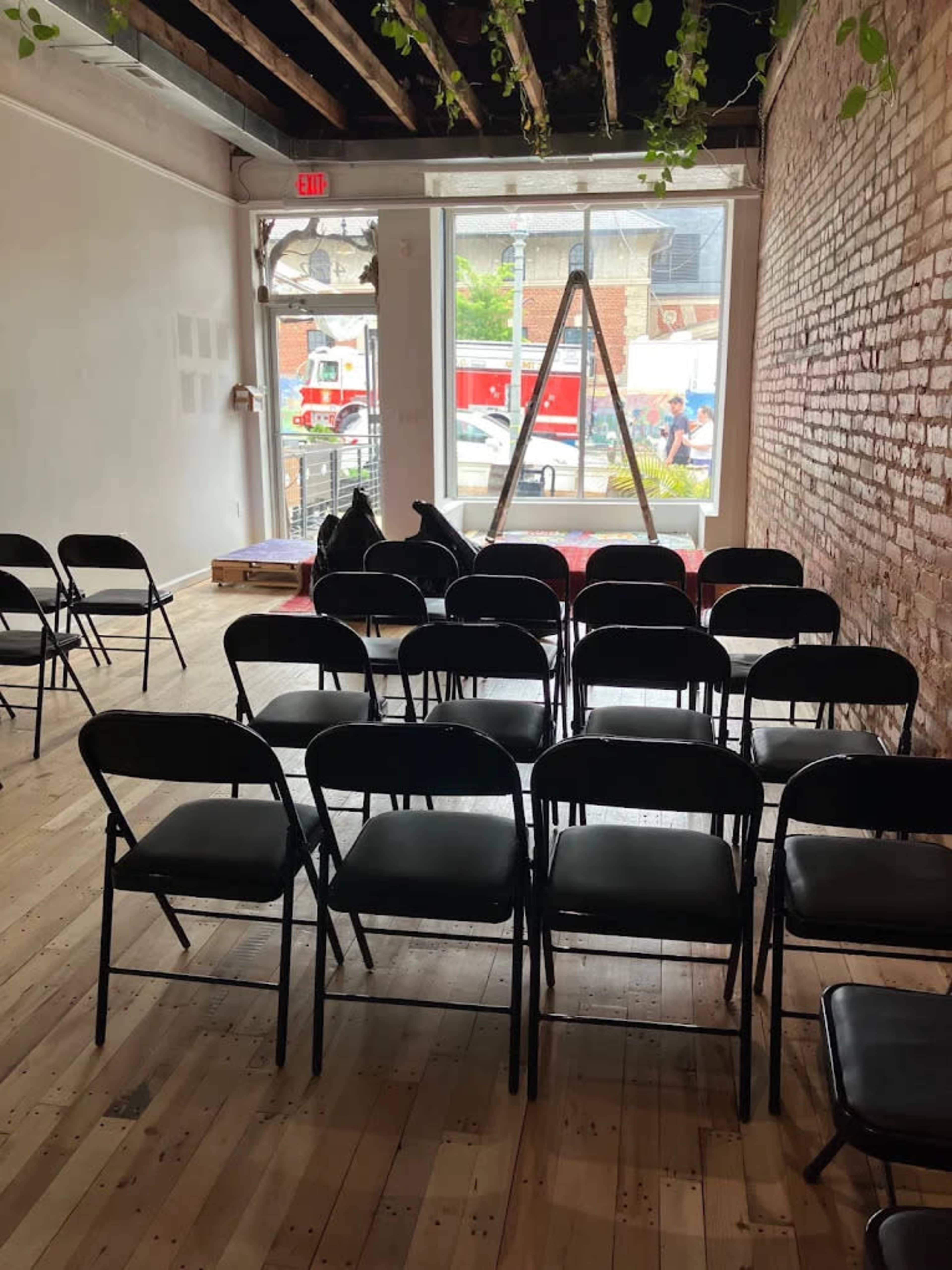The image shows a minimalist indoor space with rows of black folding chairs set up, facing a window displaying a view of the street outside.