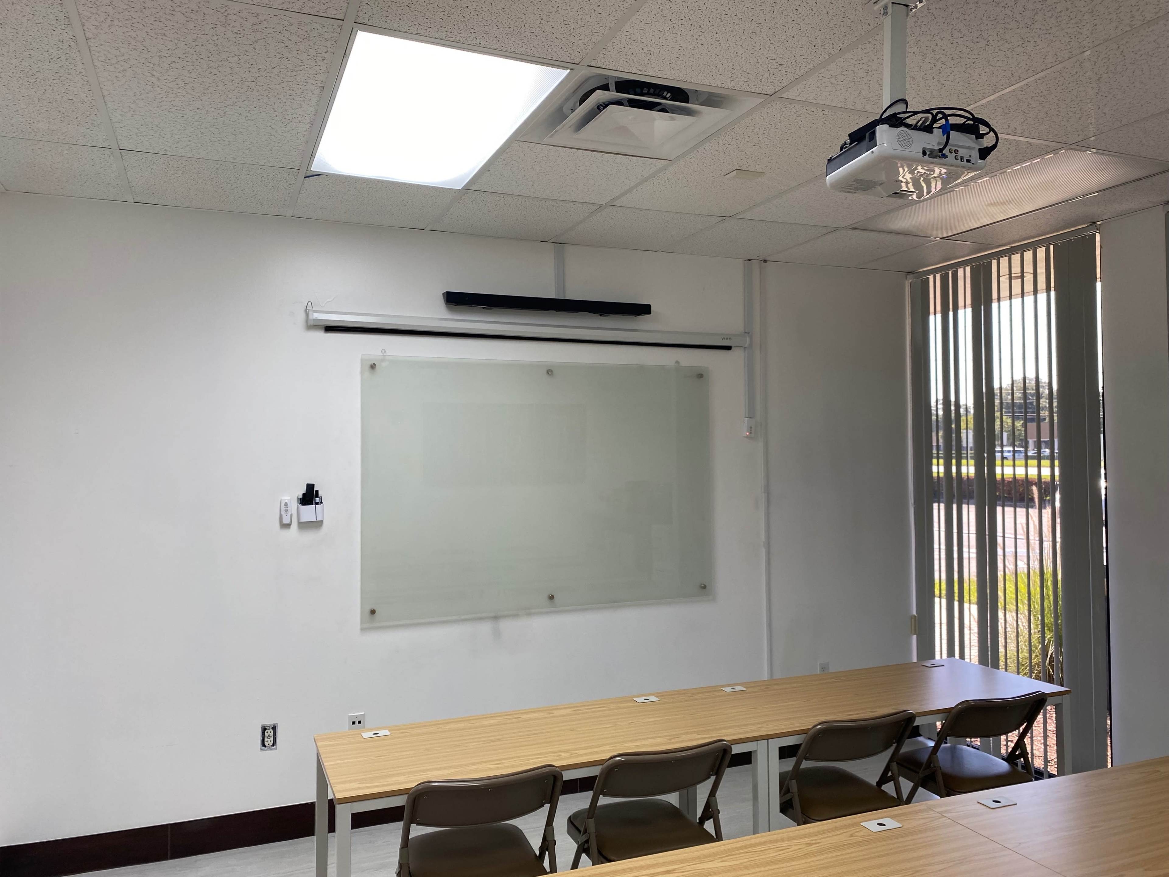A classroom setting features a long table with chairs, a white projection screen mounted on the wall, and large windows providing natural light.