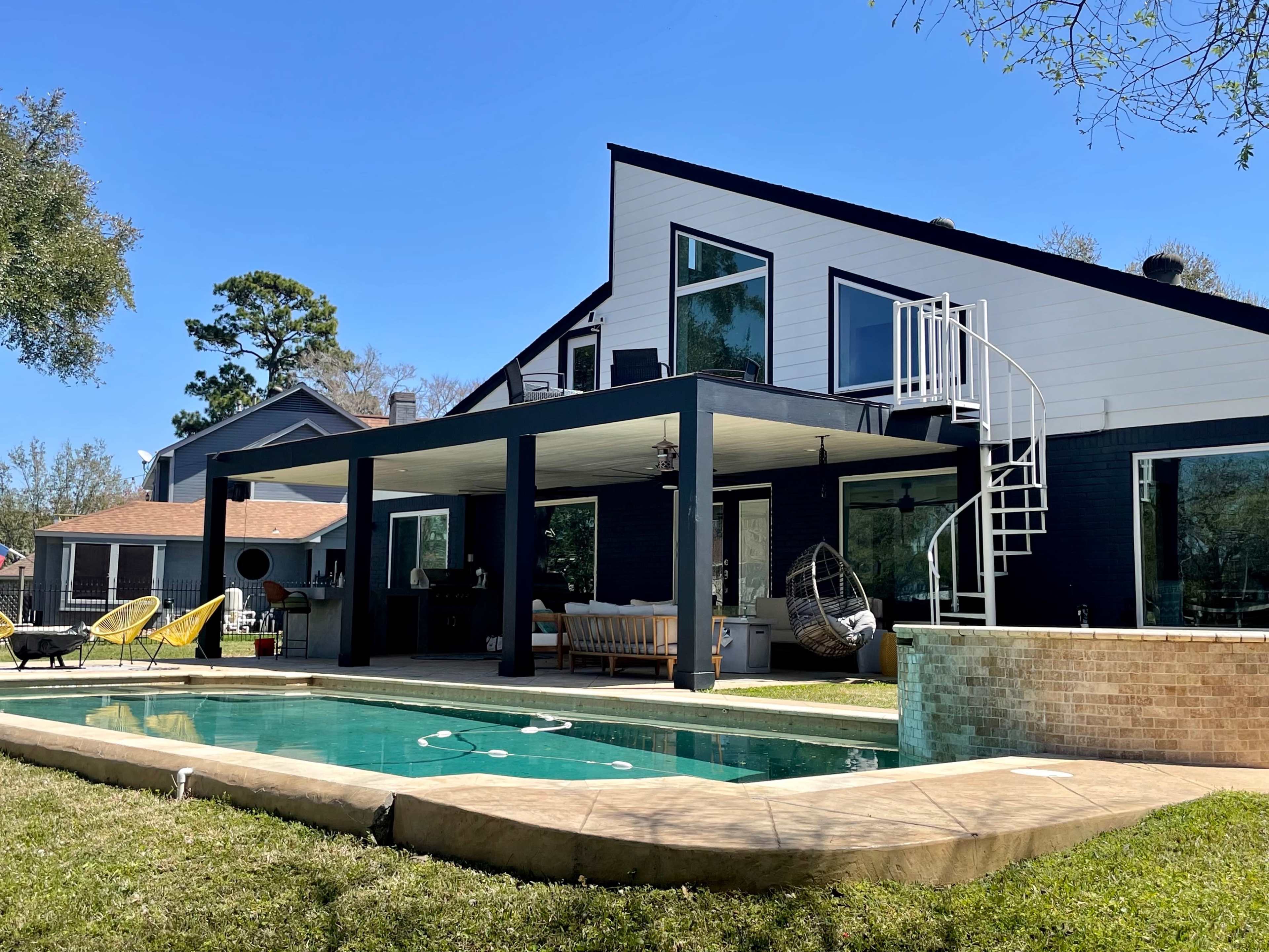 A modern home features a pool, a covered patio area with seating, and a spiral staircase leading to an upper level.
