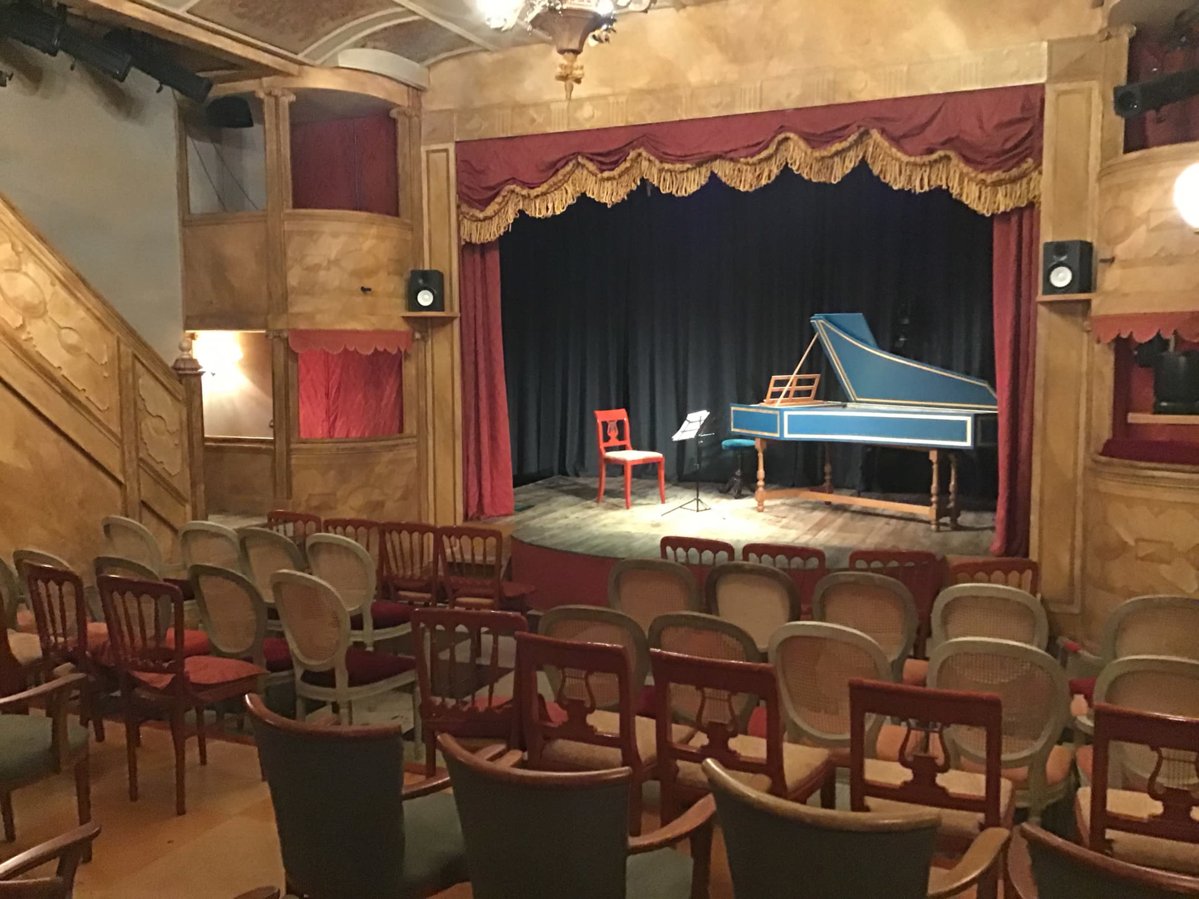 A small theater with wooden seating faces a stage adorned with red curtains and a blue harpsichord.