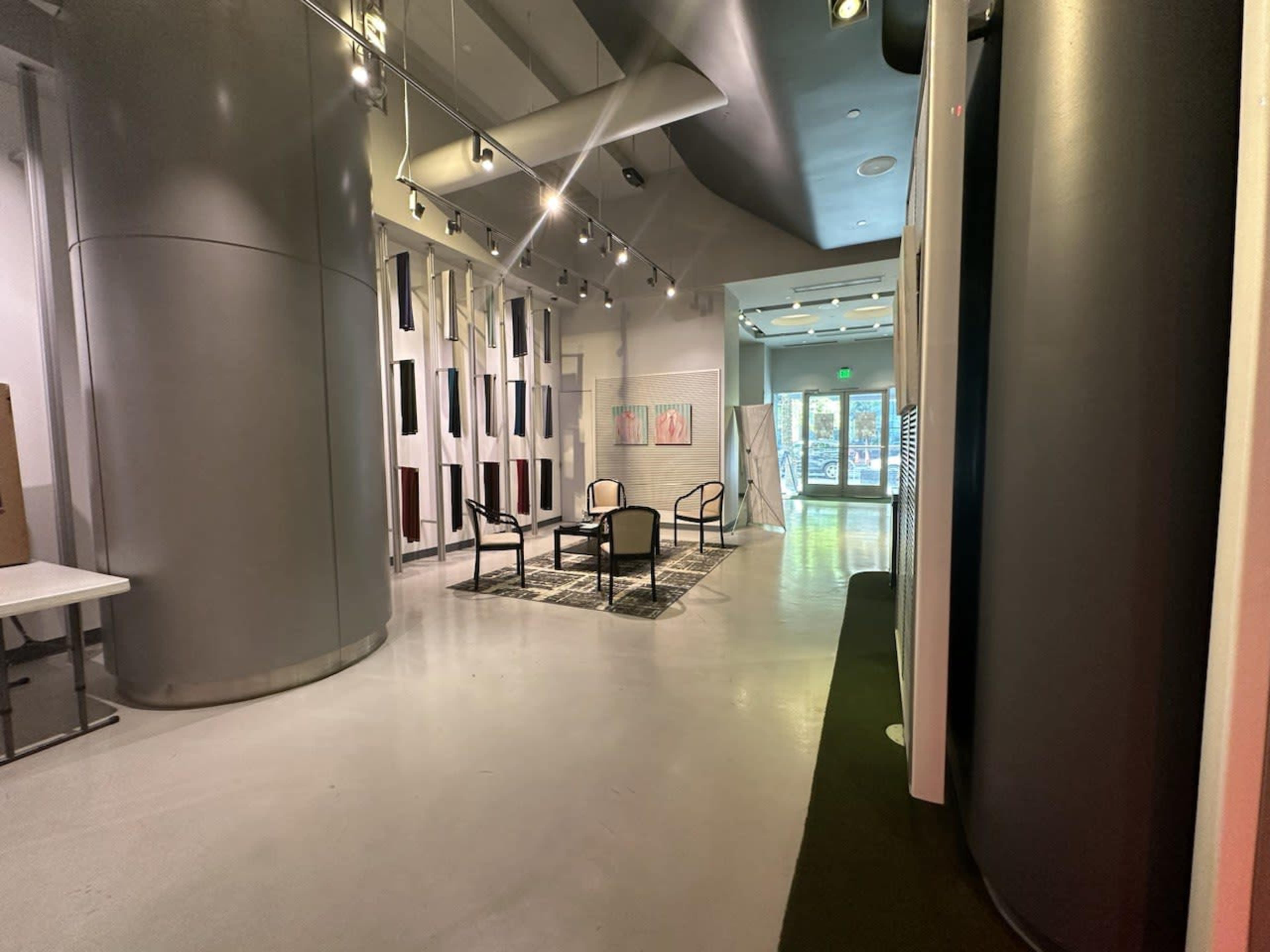 The image shows a modern, spacious interior with chairs arranged on a patterned rug, surrounded by sleek walls and ample lighting.