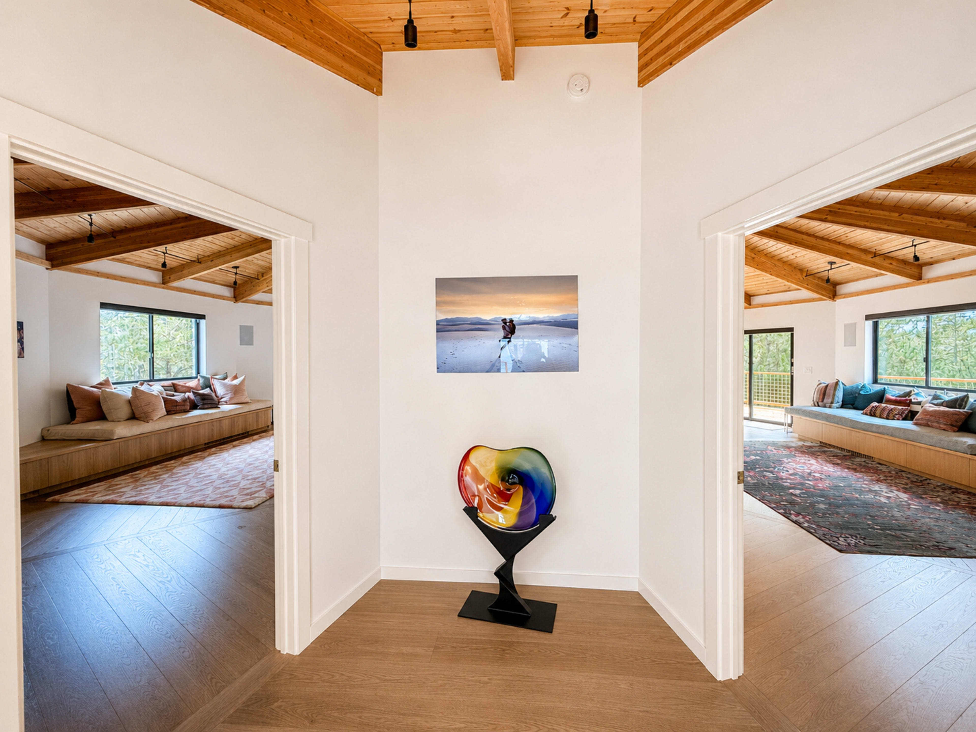 The image shows a corner of a house with two open doorways leading to different rooms, featuring a colorful sculptural piece in the foreground and large windows in the background.