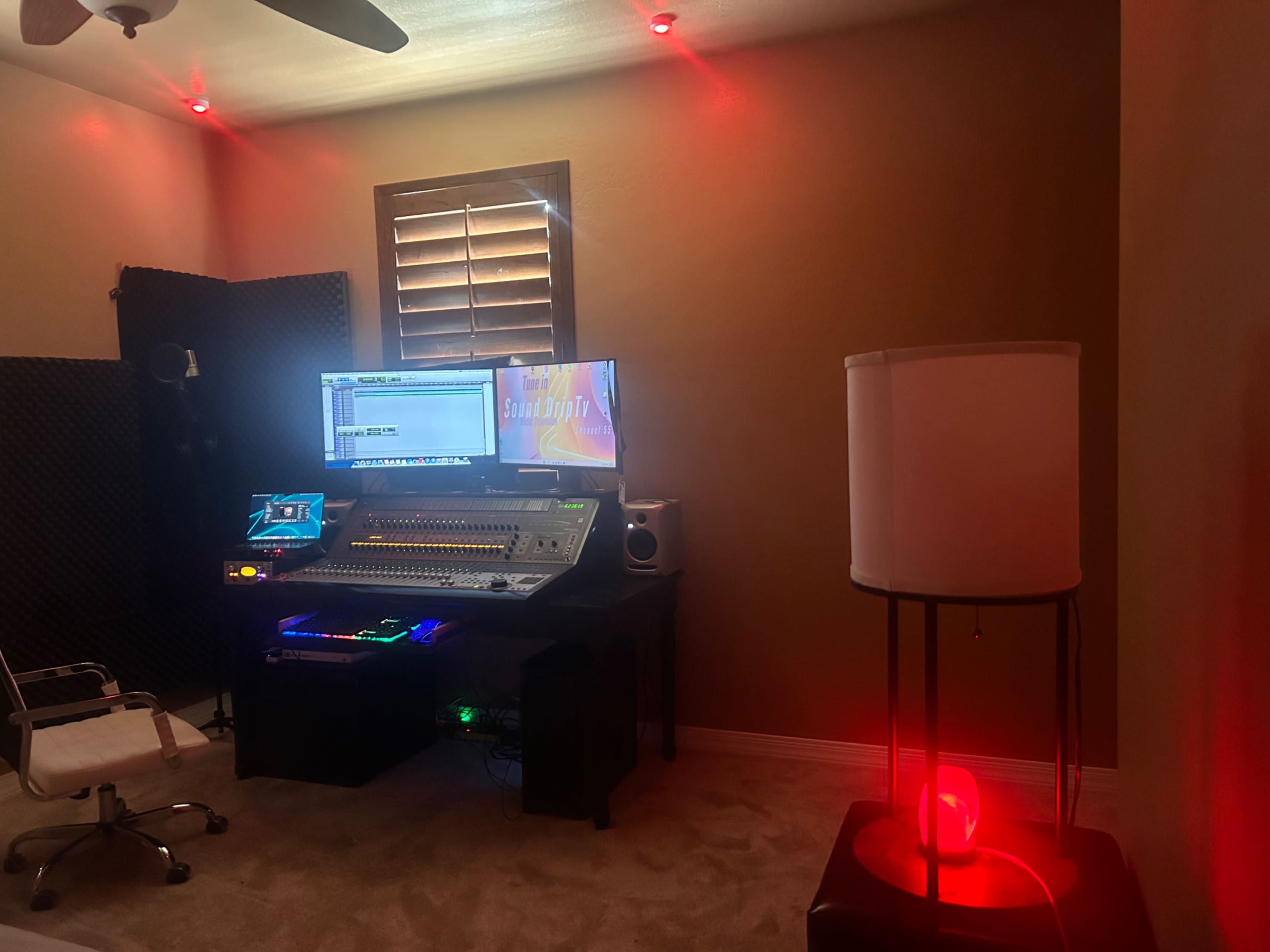 The image shows a recording studio featuring a large mixing console, a computer monitor, and ambient red lighting, with soundproofing panels on the walls.