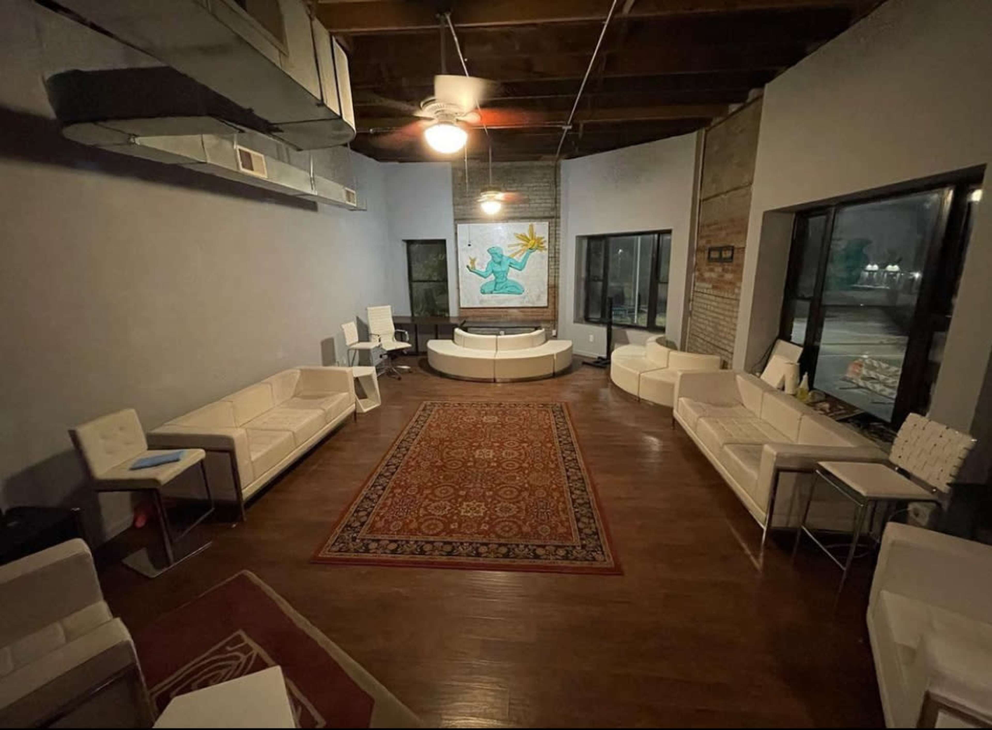A spacious room with white furniture arranged around a central circular table, featuring a patterned rug on the wooden floor and a large artwork on the wall.