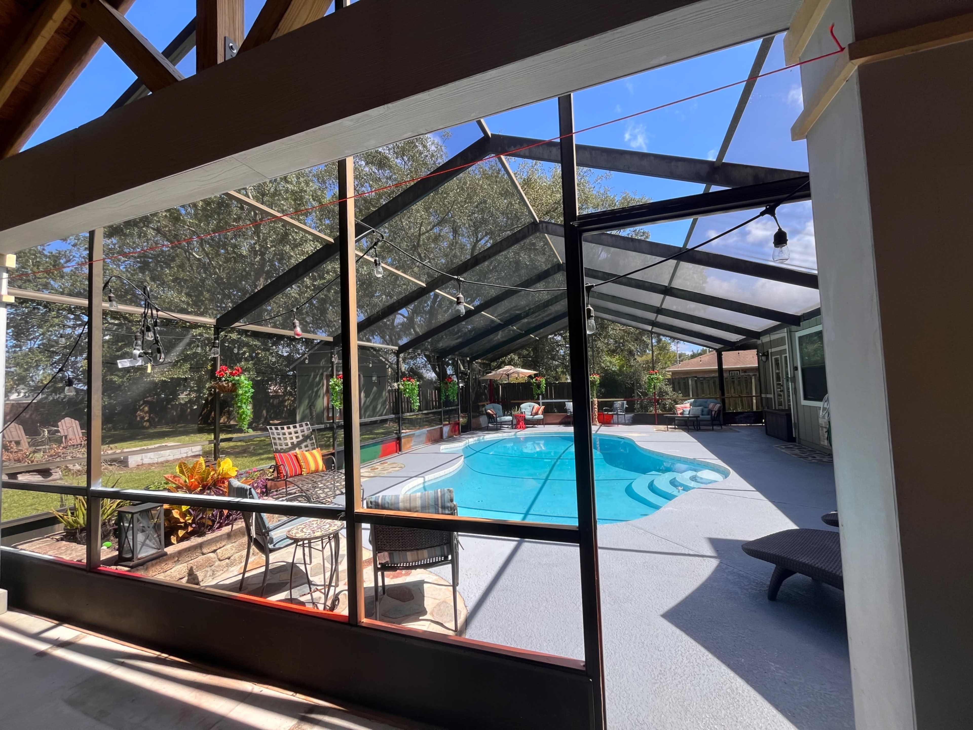 The image shows a screened-in pool area with a clear blue pool, surrounded by patio furniture and landscaping under a bright sky.