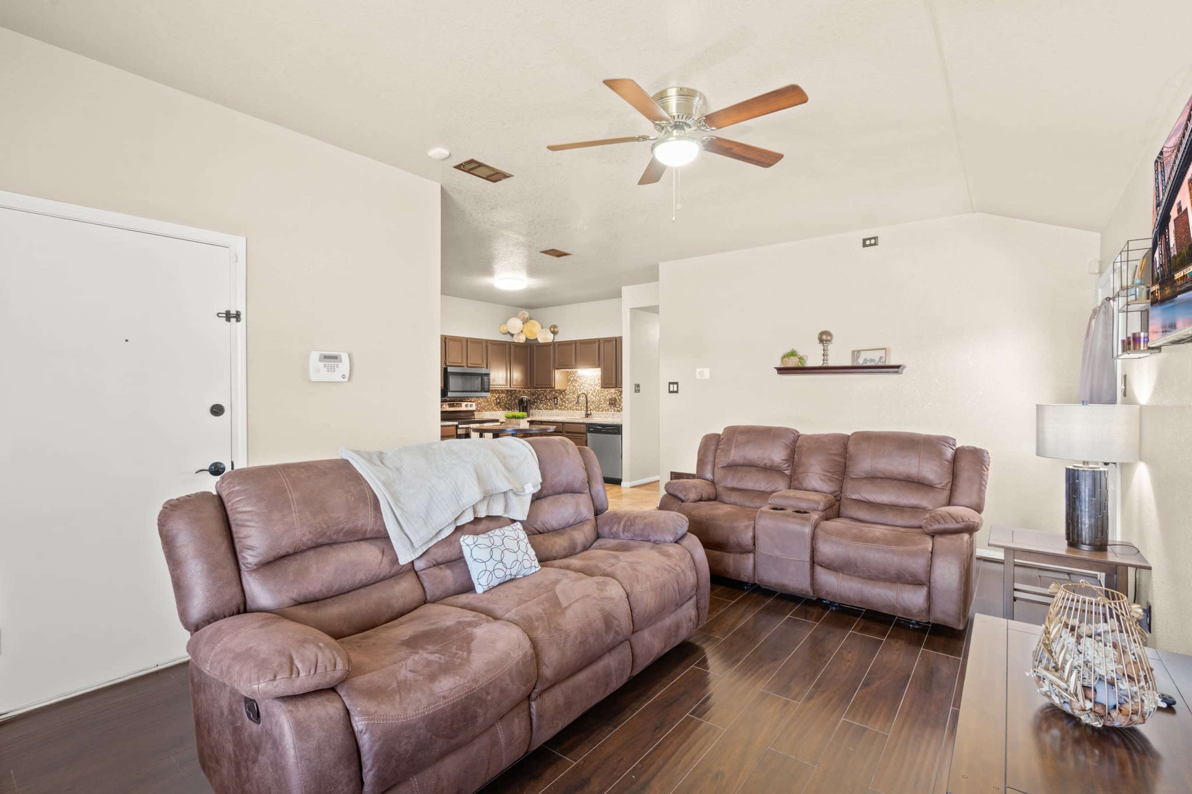 A living room with brown reclining sofas, a ceiling fan, and a view of a kitchen in the background.