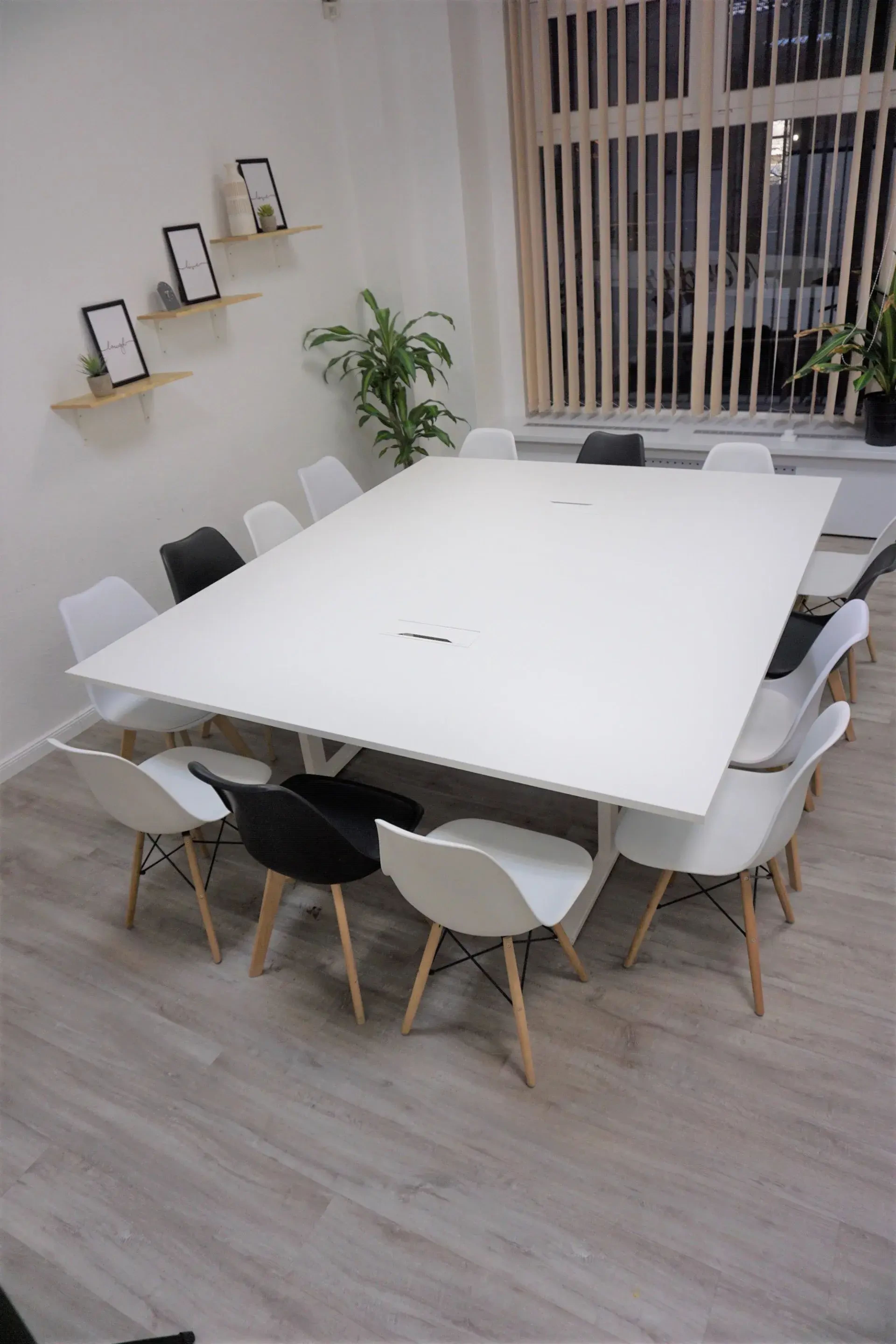 A large white conference table surrounded by black and white chairs is arranged in a well-lit room with plants and framed pictures on the walls.
