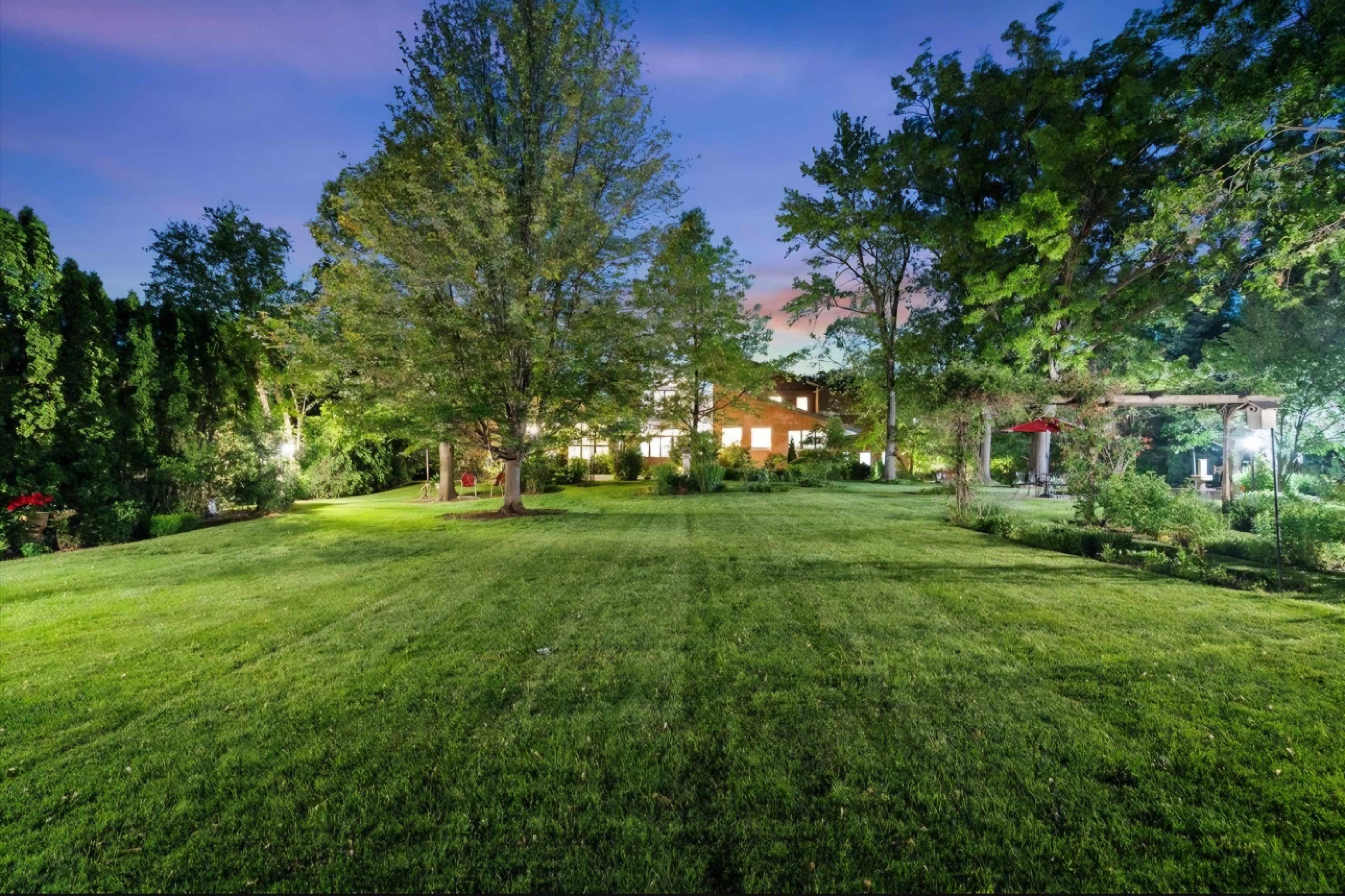 The image showcases a well-manicured lawn surrounded by trees and a house, with soft lighting illuminating the area during twilight.