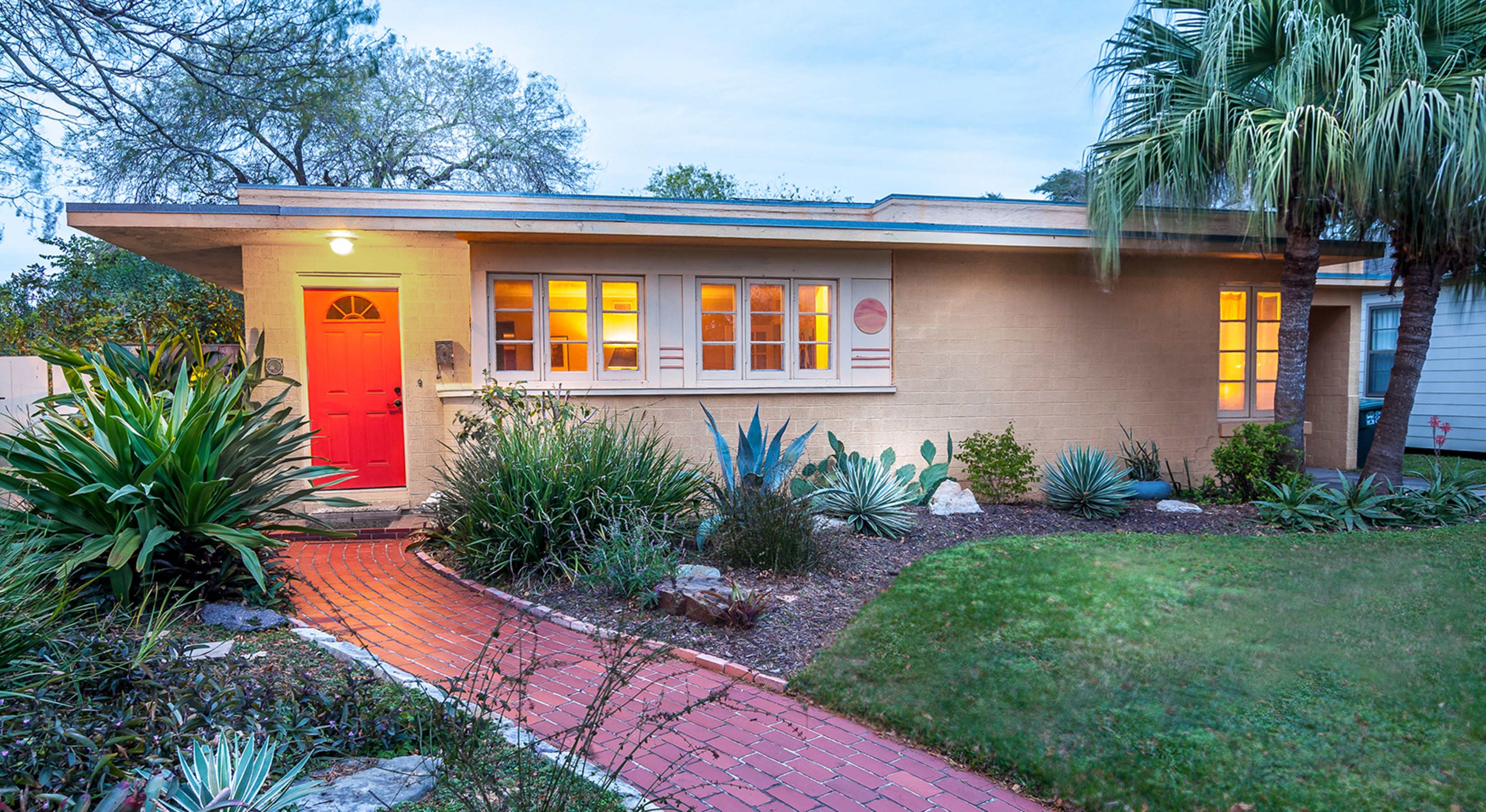 A modern house with a red door sits at the end of a brick pathway, surrounded by various shrubs and palm trees.