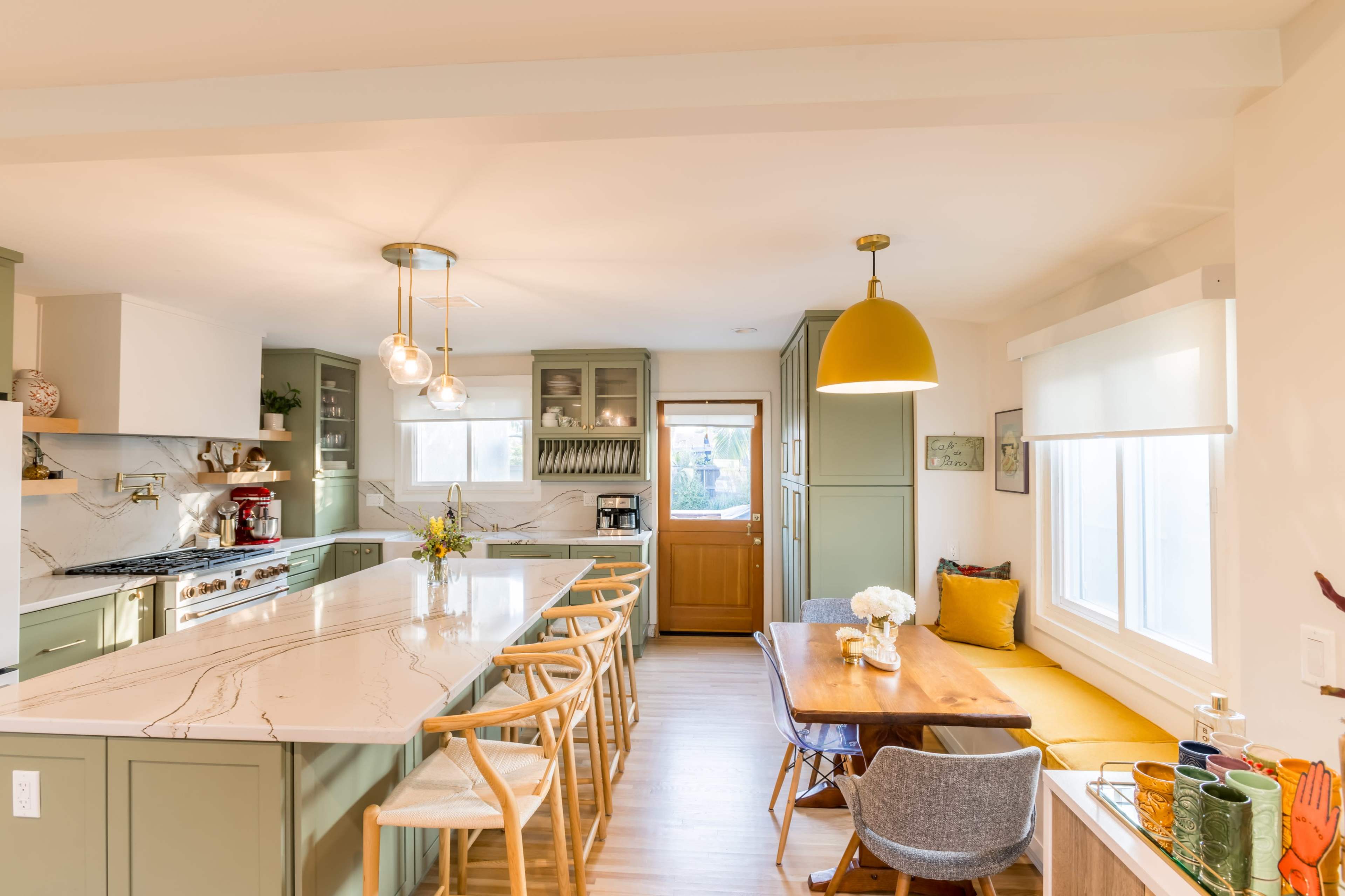 A modern kitchen with green cabinetry, a large island with a marble countertop, a wooden dining table, and a yellow pendant light.