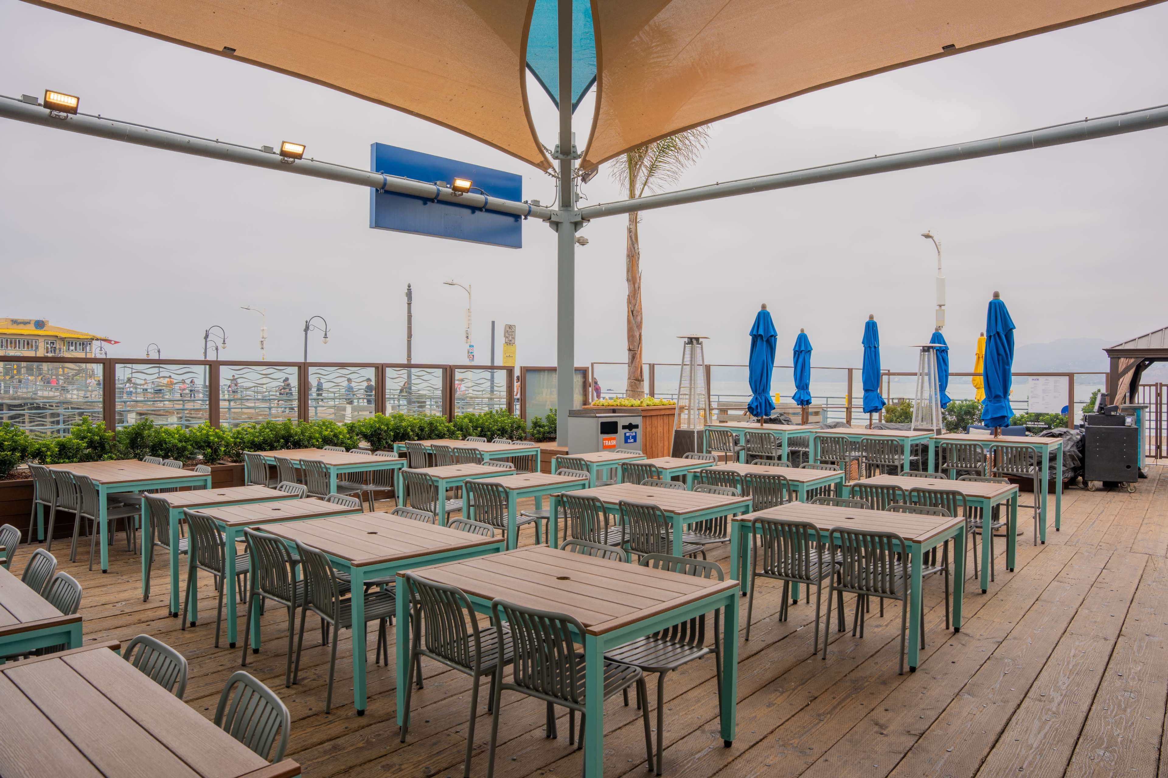 An outdoor dining area with wooden tables and chairs is set up under a canopy, facing a view of the ocean.