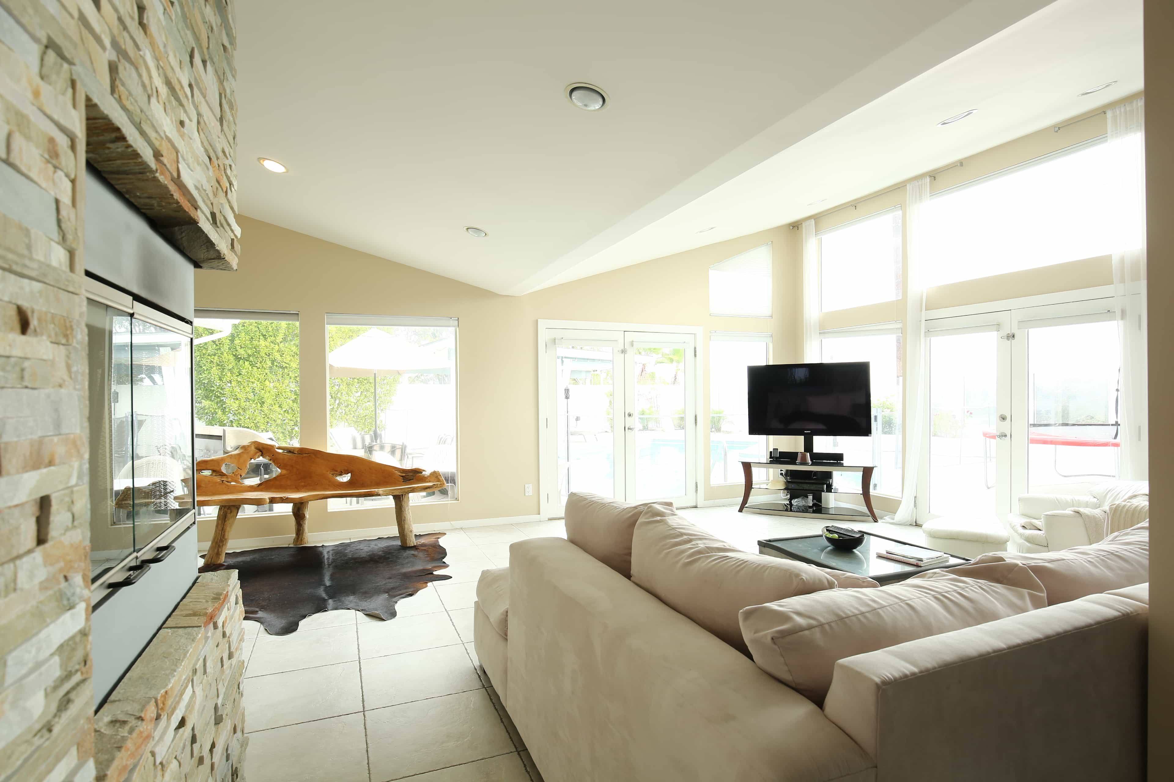 A modern living room with a stone fireplace, a large sofa, and floor-to-ceiling windows overlooking a pool area.