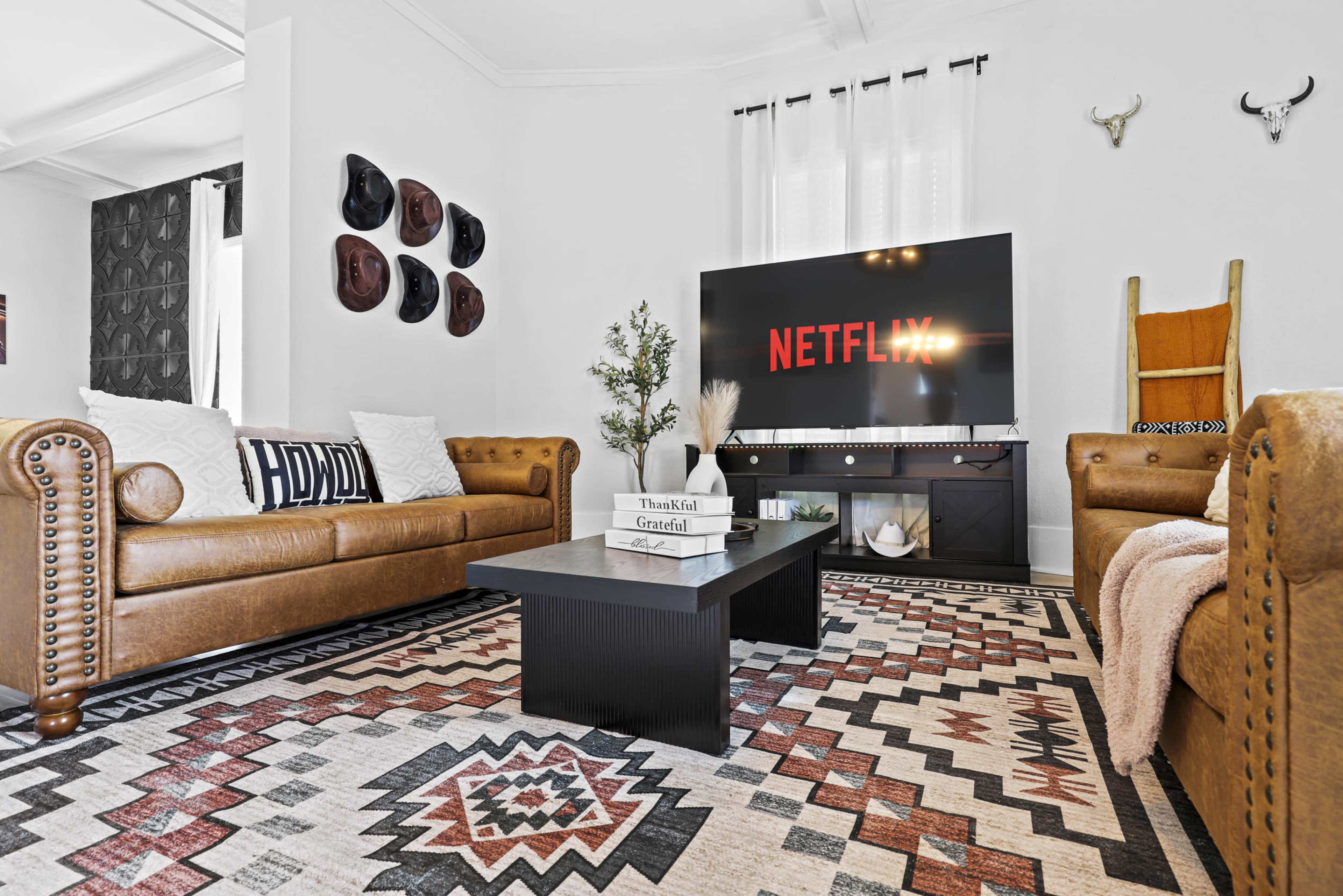 A living room features two brown leather couches, a black coffee table, and a television displaying the Netflix logo, with decorative wall hangings and a patterned rug.