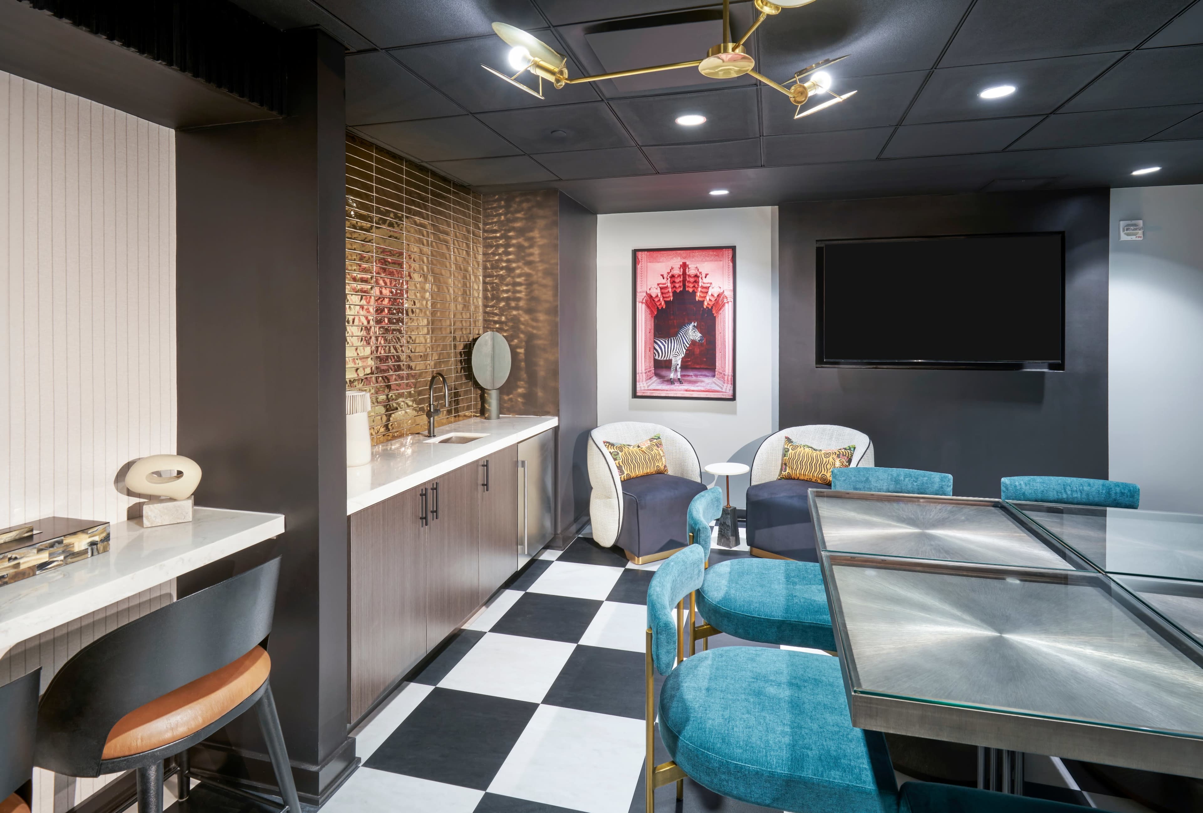 The image shows a modern lounge area with a sleek bar counter, plush seating, and a television mounted on the wall, featuring black and white checkered flooring and artistic decor.