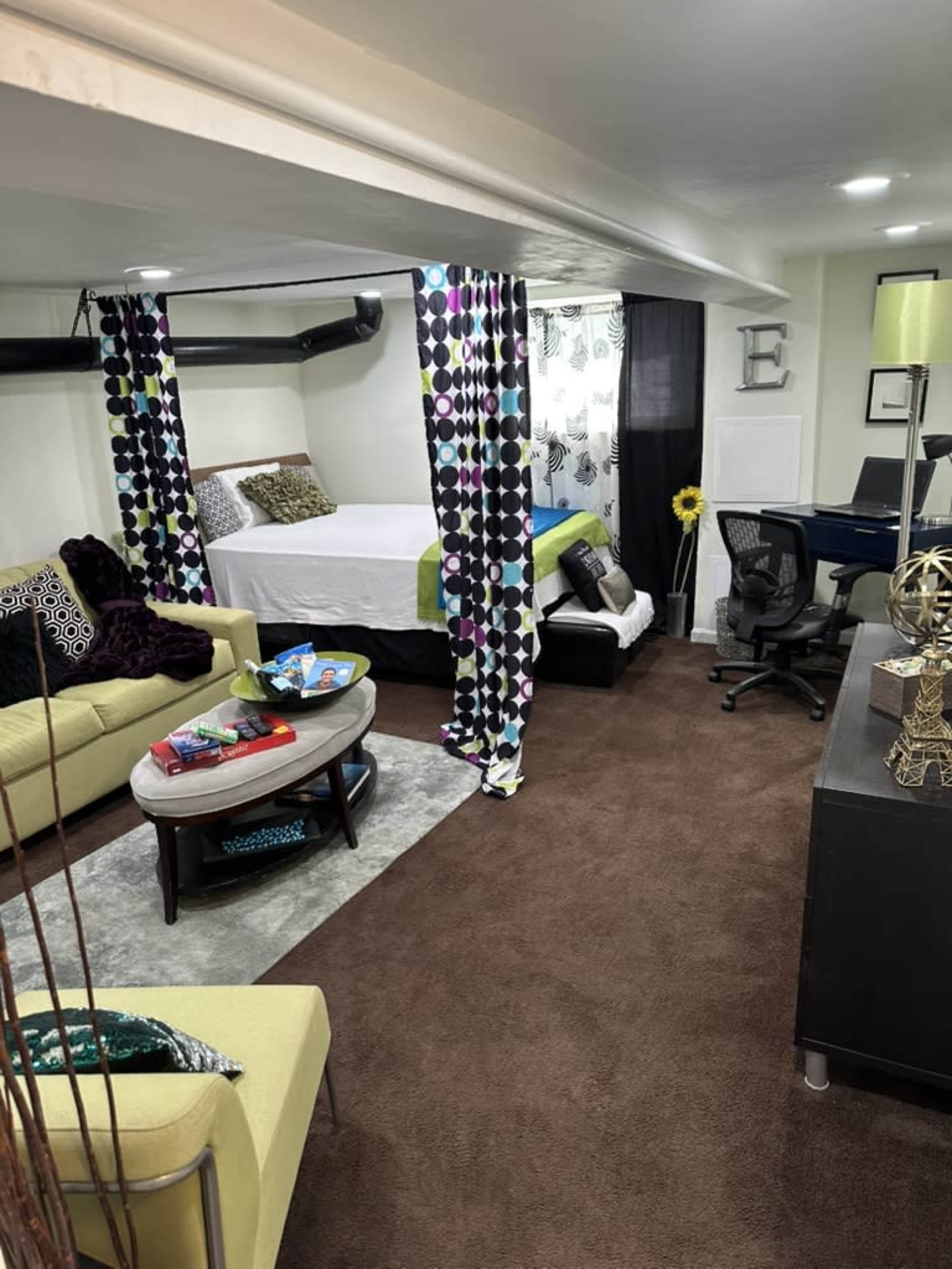 The image shows a cozy basement room with a bed, seating area, desk, and decorative curtains separating spaces.