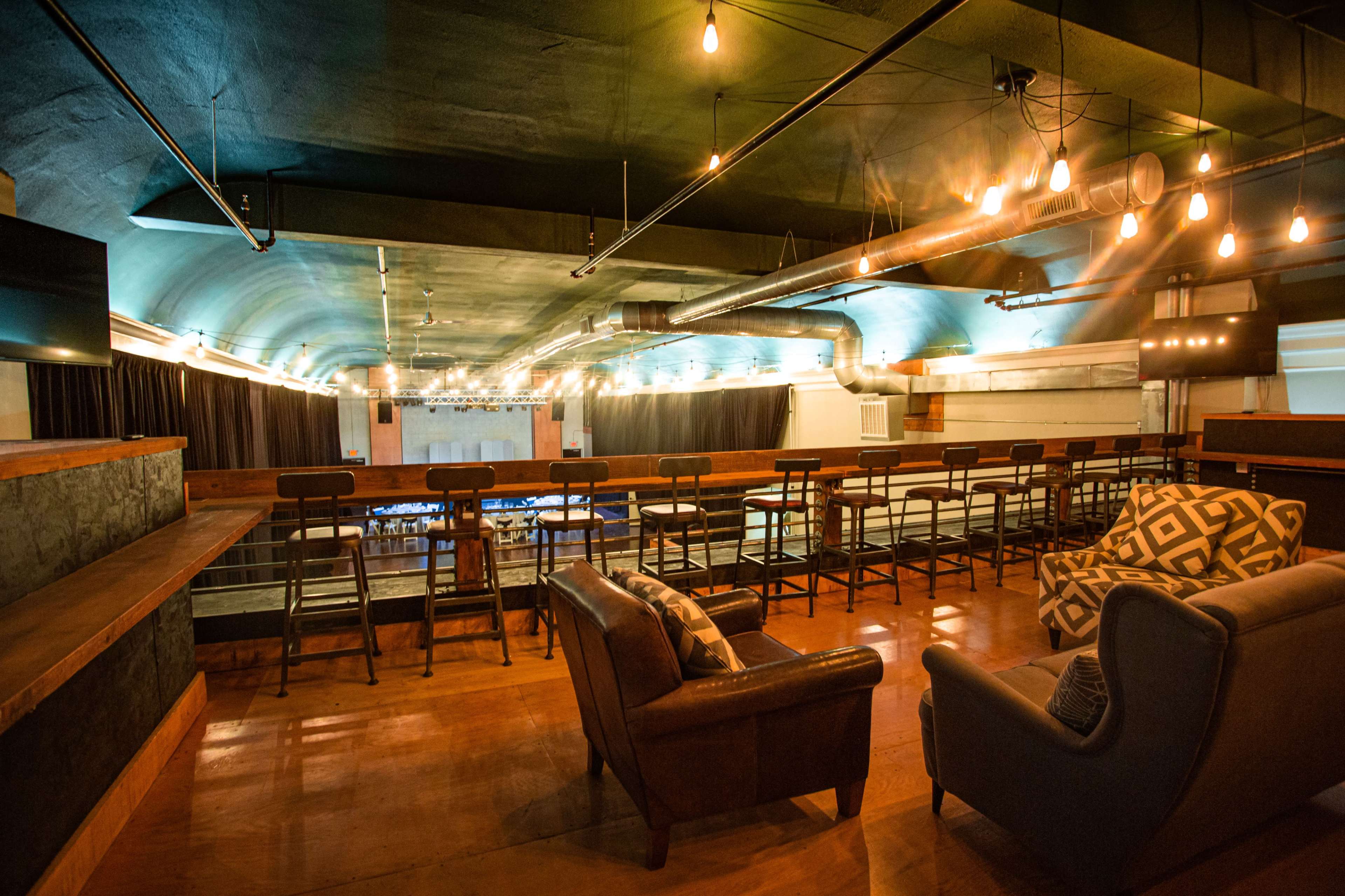 The image shows a spacious, modern interior of a venue with a bar area, seating arrangements, and soft lighting fixtures hanging from the ceiling.