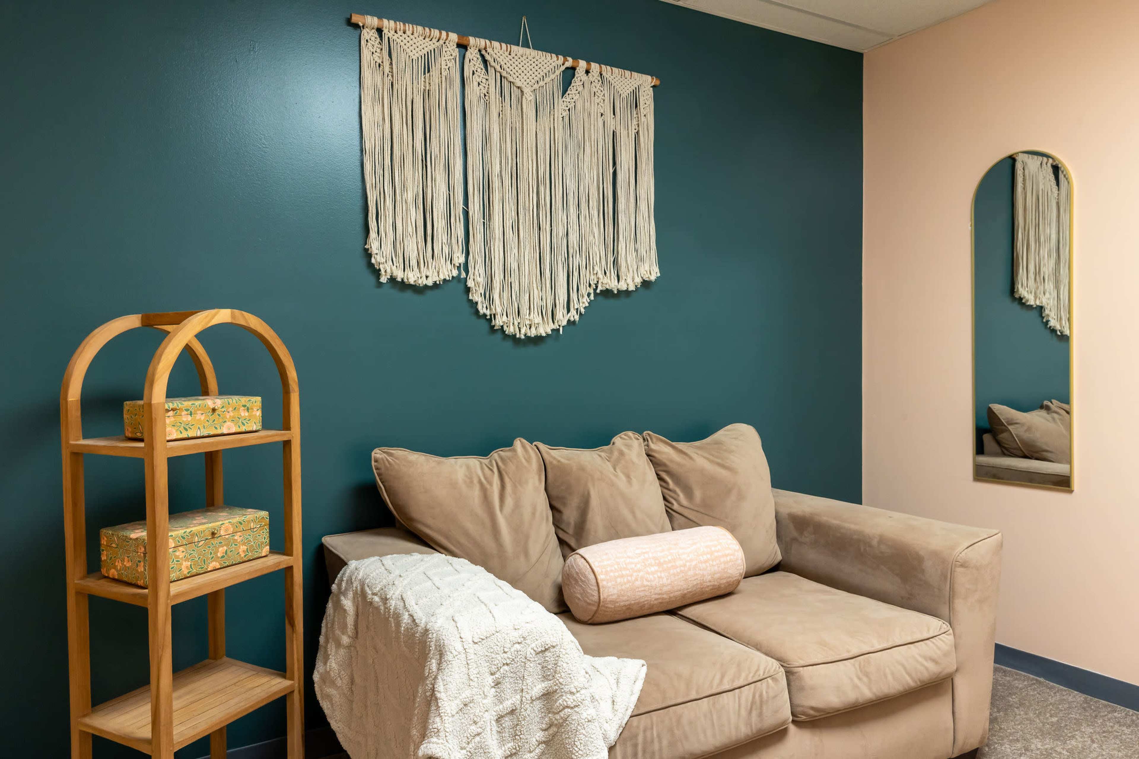 The image shows a cozy living space featuring a brown sofa, a decorative wall hanging, a wooden shelf with patterned boxes, and a mirror on the adjacent wall.