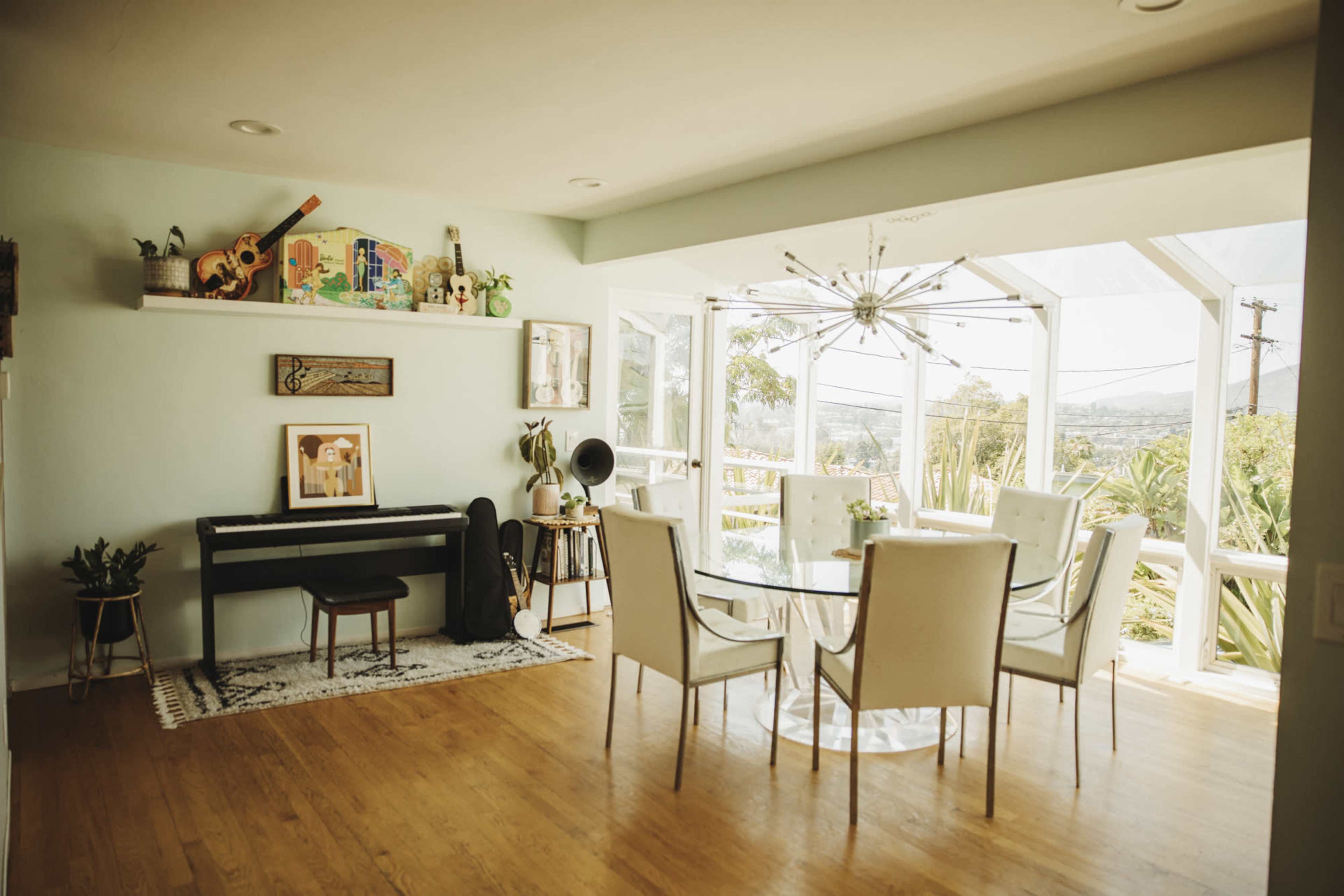A light-filled dining area features a glass table surrounded by white chairs, with a piano and decorative elements on the walls.