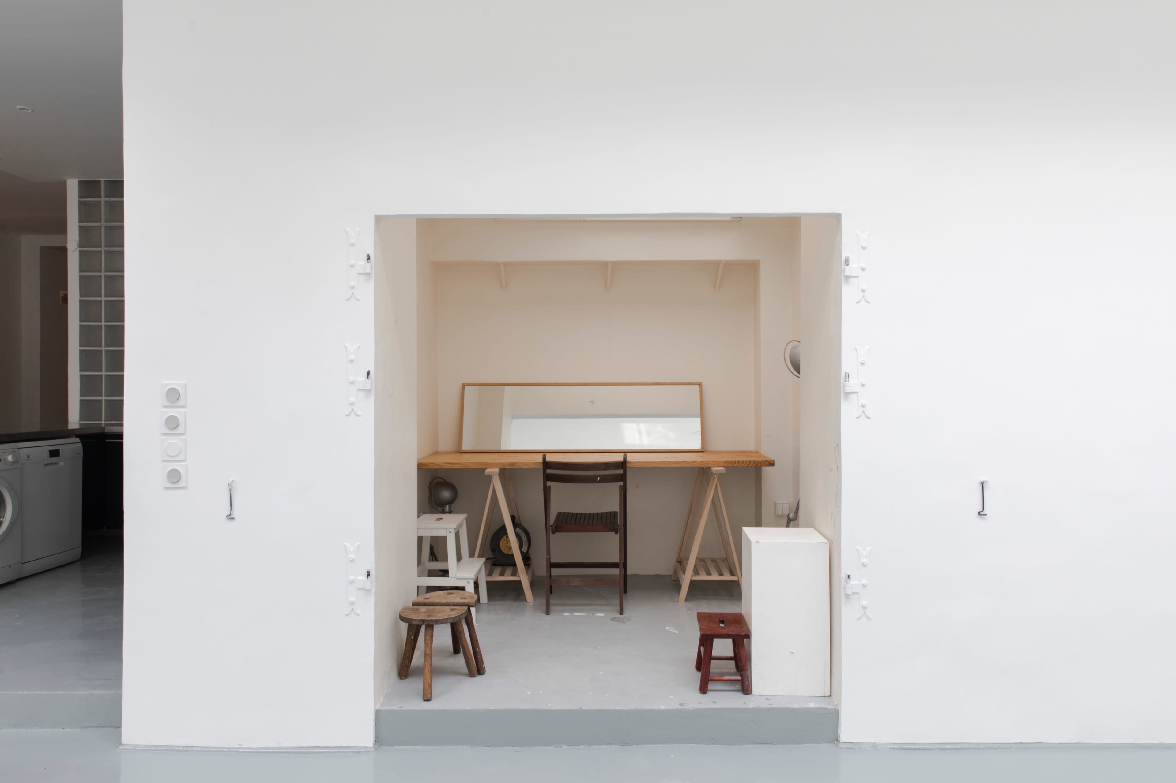 A minimalist room with a wooden table, a large mirror, and several stools arranged in a recessed area.