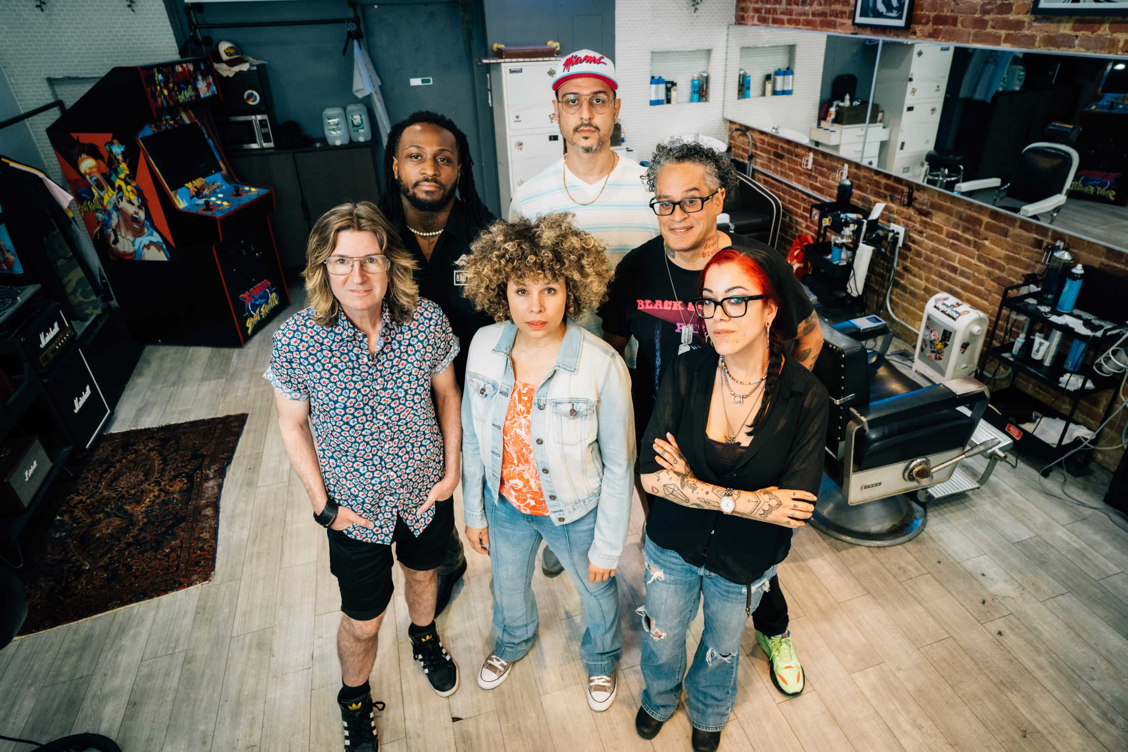 A group of six people stands together inside a barbershop, with vintage arcade machines and barber chairs visible in the background.