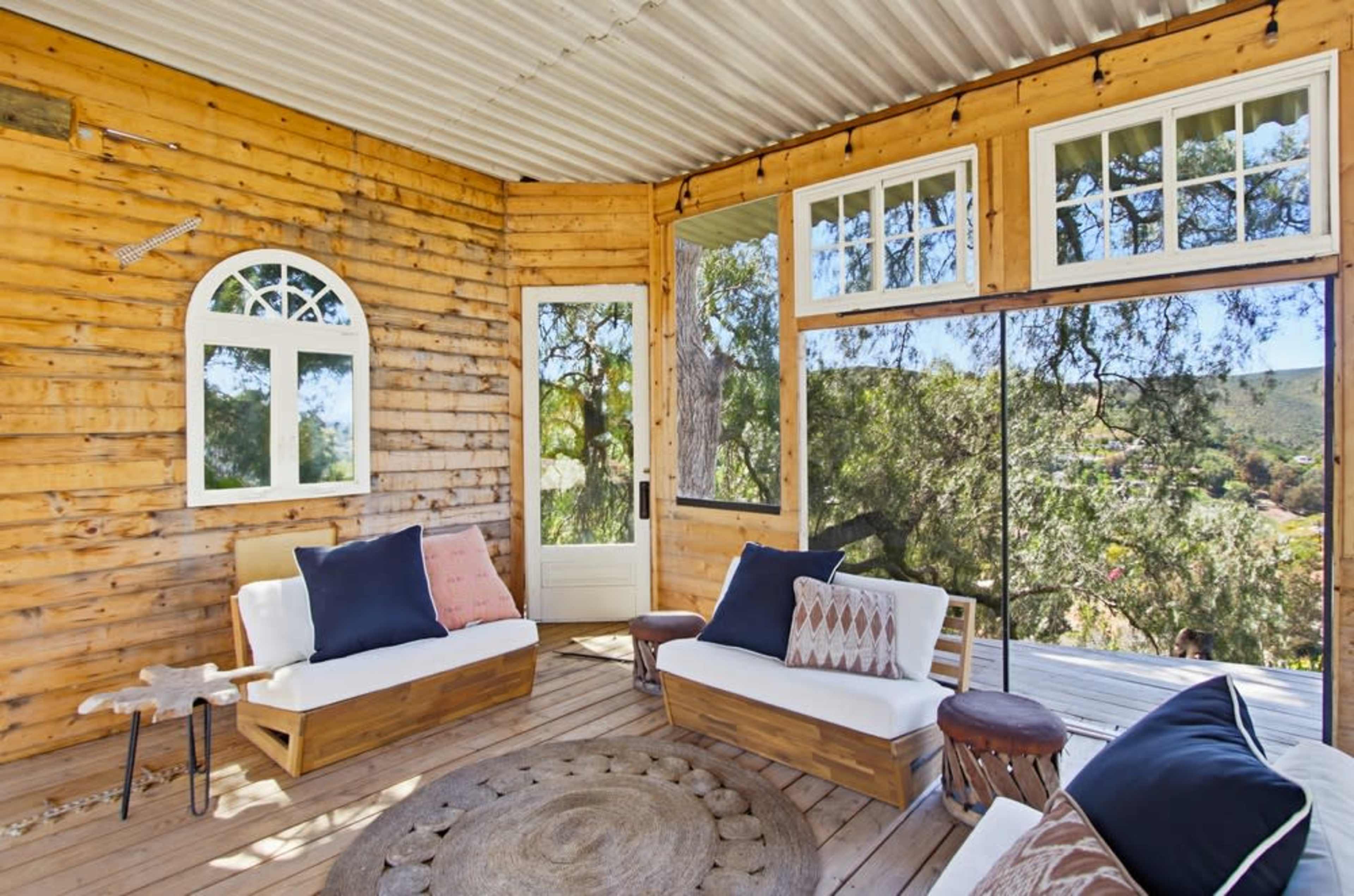The image shows a wooden porch with two white sofas, a circular rug, and large windows overlooking a scenic view.
