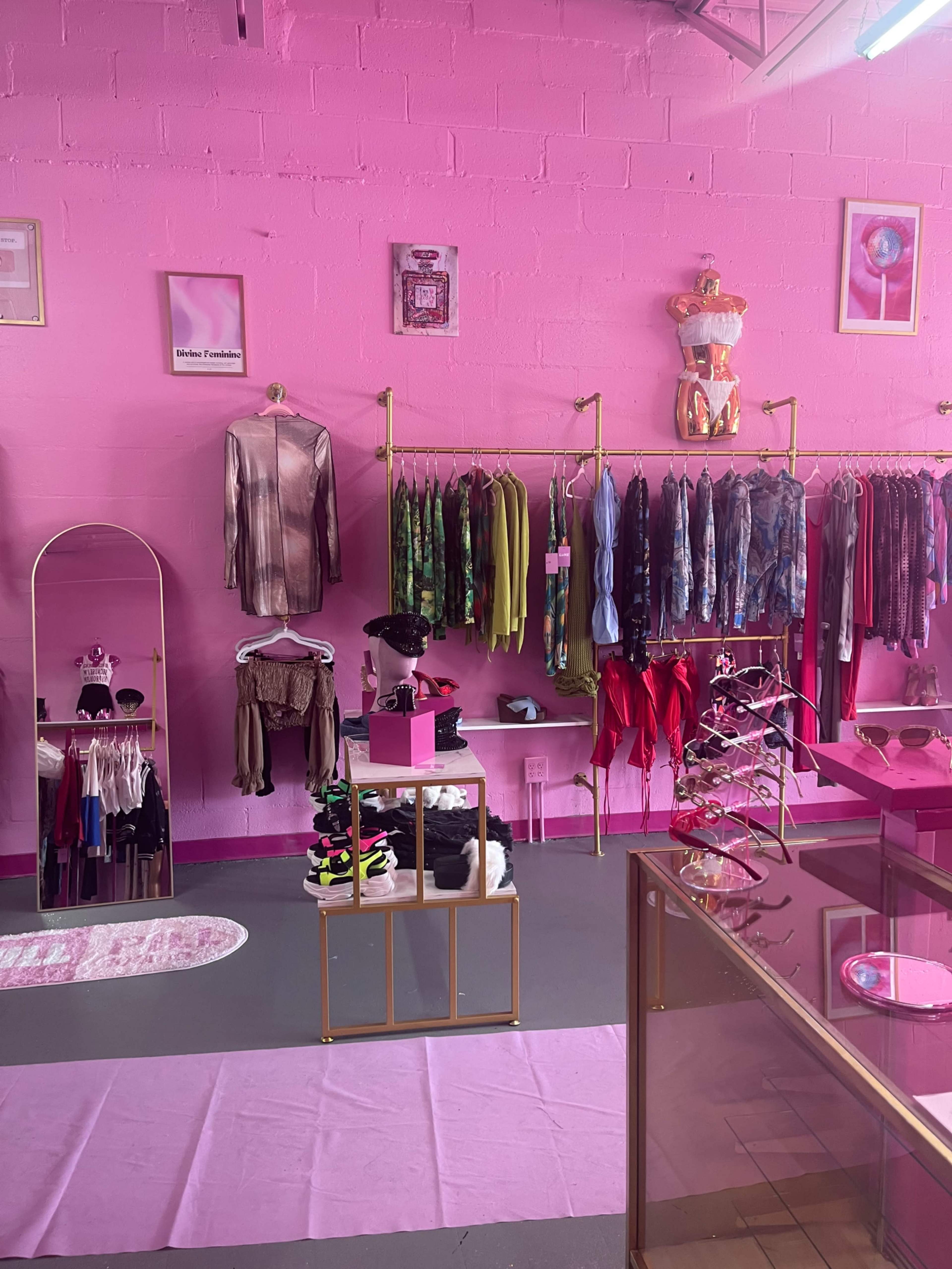 The image shows a boutique interior with vibrant pink walls, displaying clothing racks filled with colorful garments, accessories on display, and a central pedestal featuring shoes and sunglasses.