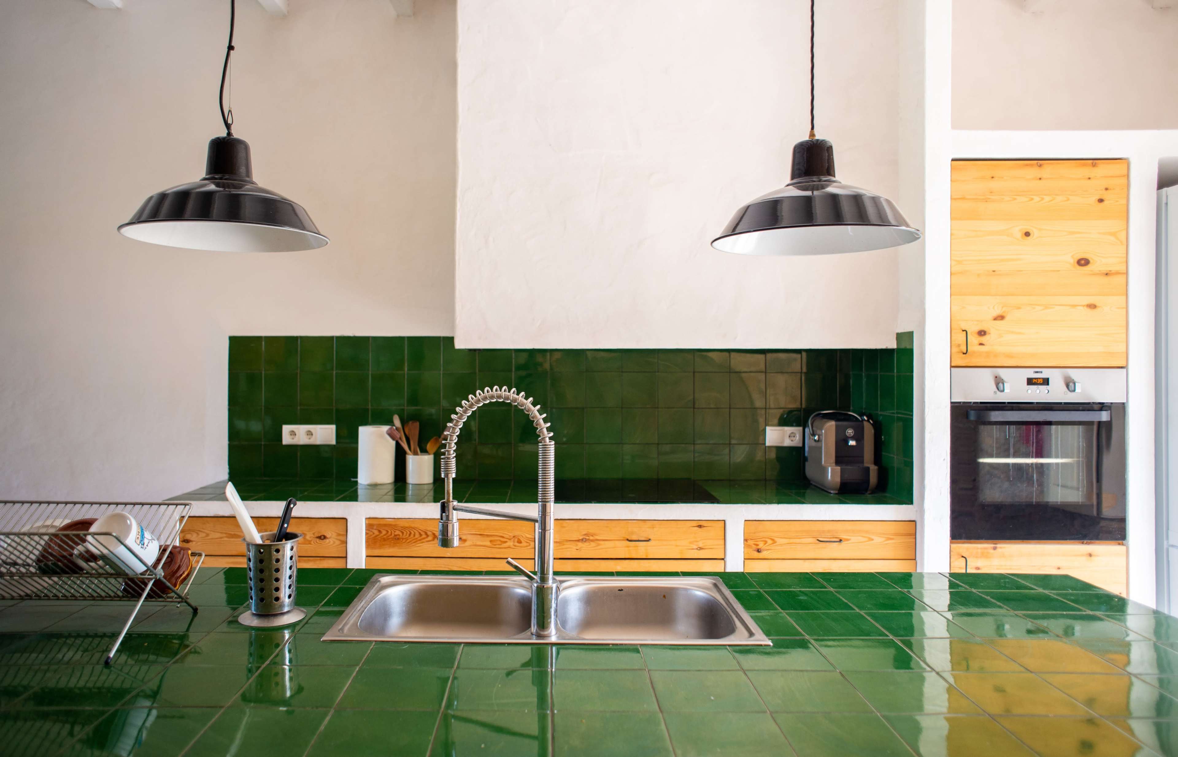 A modern kitchen features a green tiled countertop with a stainless steel sink, wooden cabinets, and two black pendant lights.