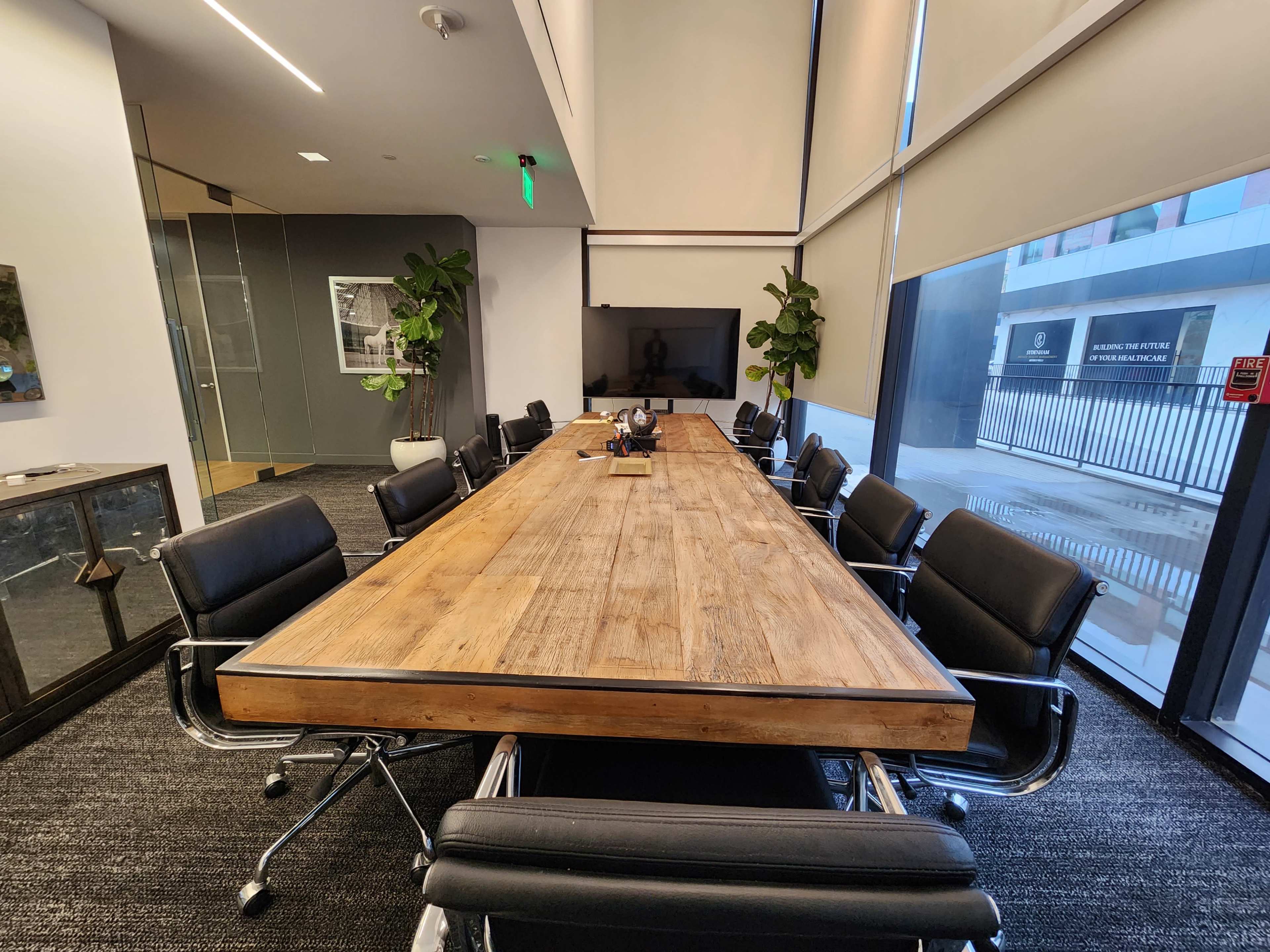A long wooden conference table surrounded by black leather chairs is set up in a modern meeting room with large windows and indoor plants.