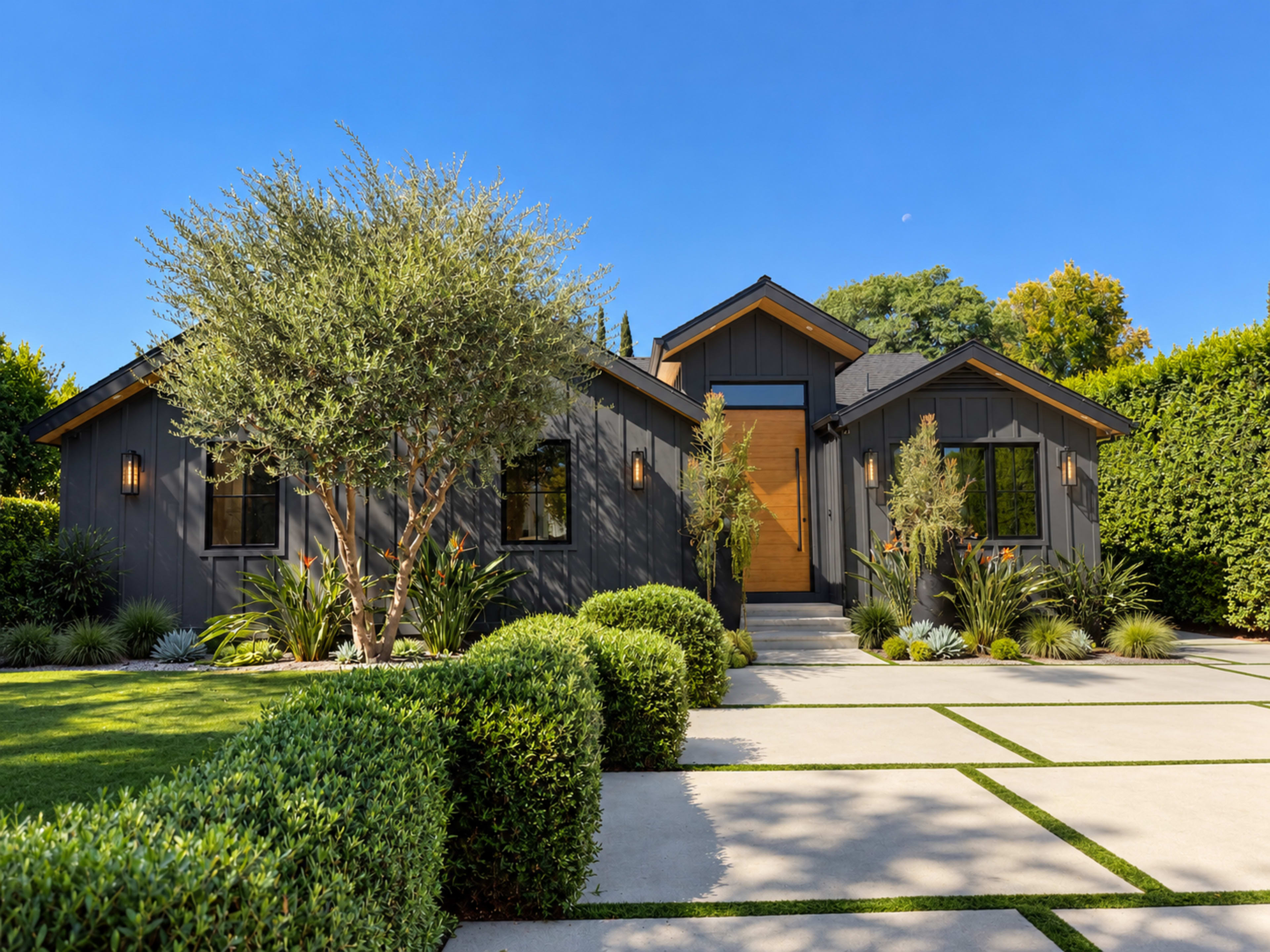 The image shows a modern one-story house with a wooden front door, surrounded by neatly landscaped greenery and a paved driveway.