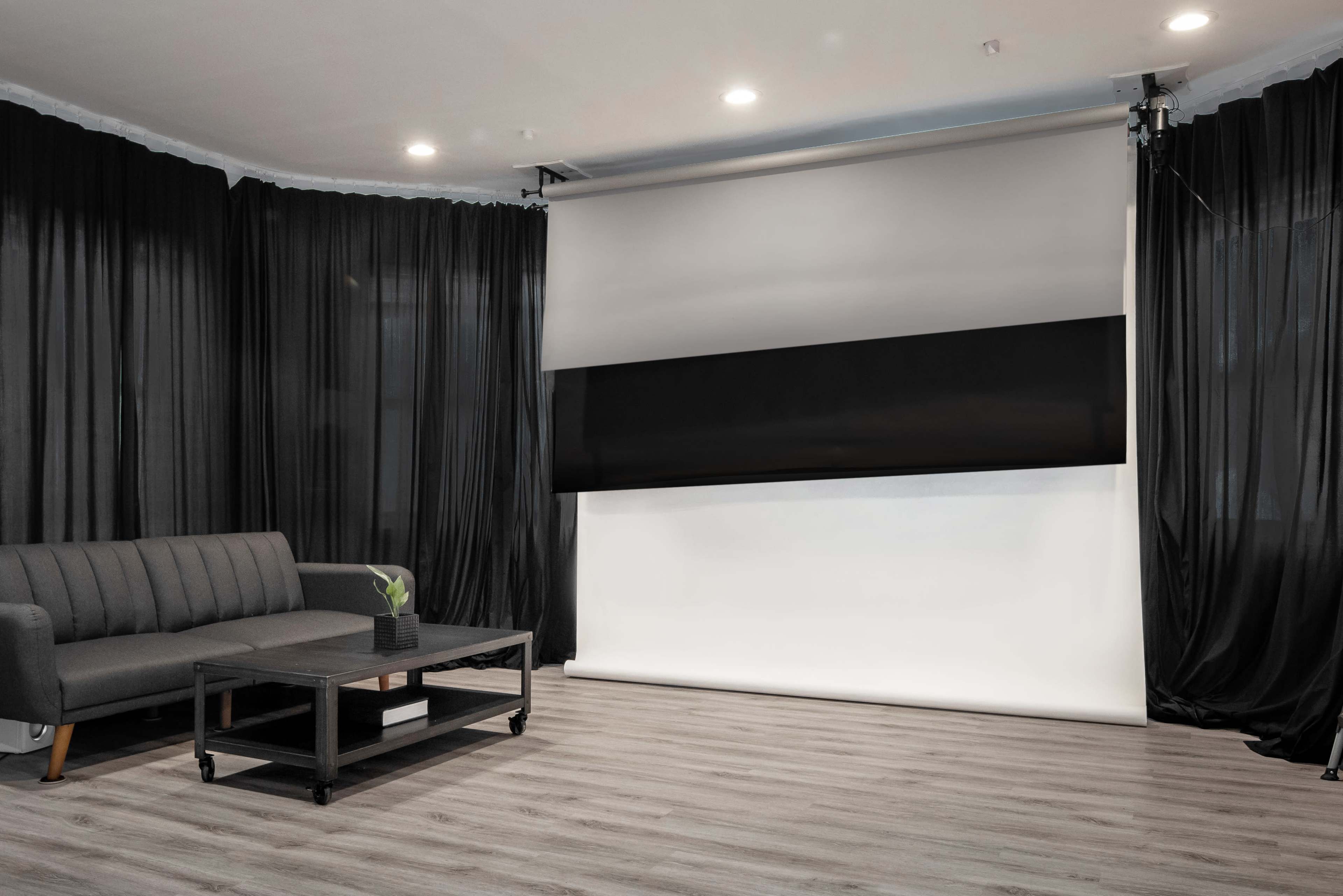 A modern living room features a gray sofa, a black coffee table, and a large retractable screen set against a backdrop of dark curtains.