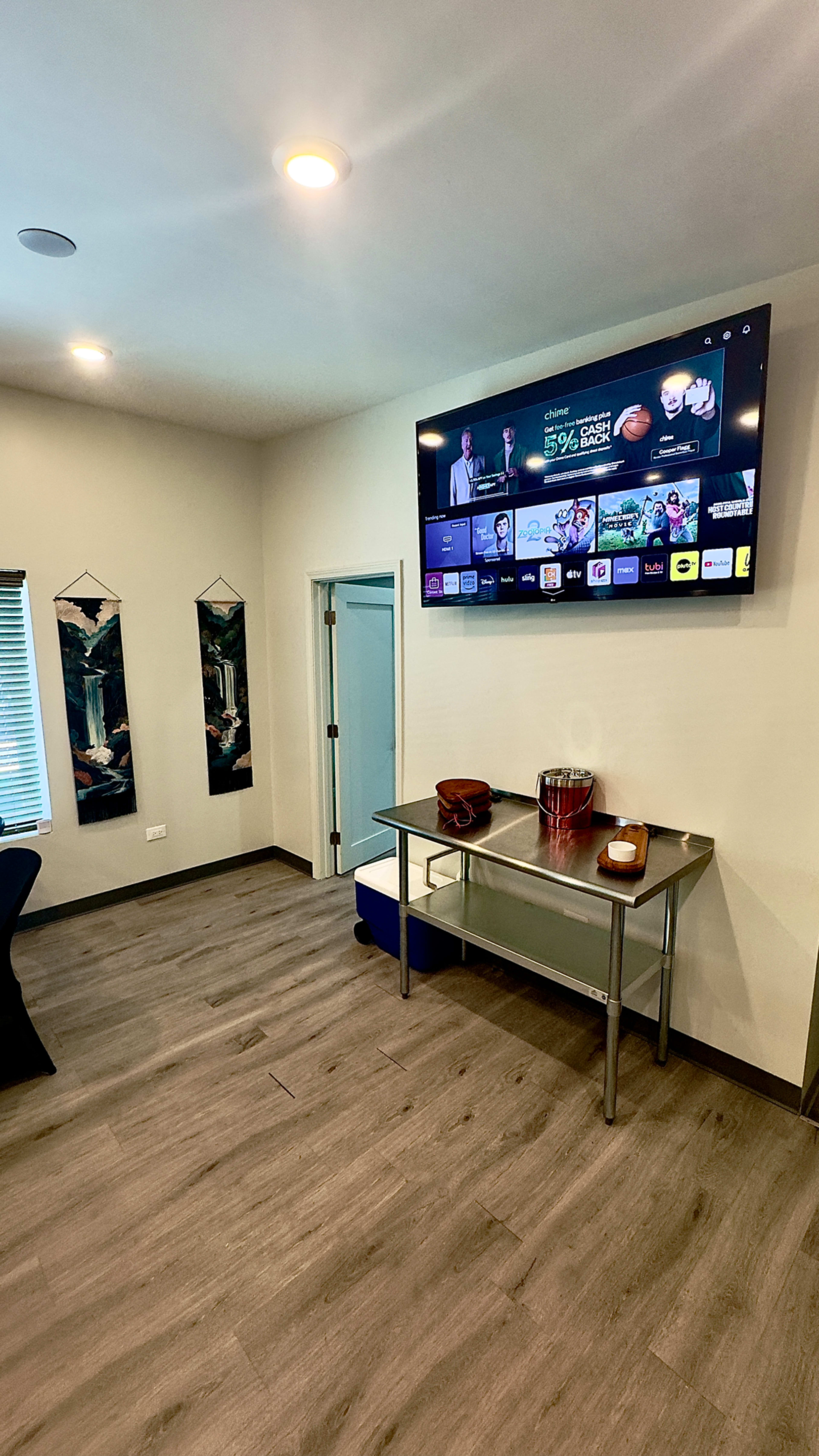 A modern room features a wall-mounted television displaying a streaming interface above a stainless steel table with various items, including a cooler and a bowl.